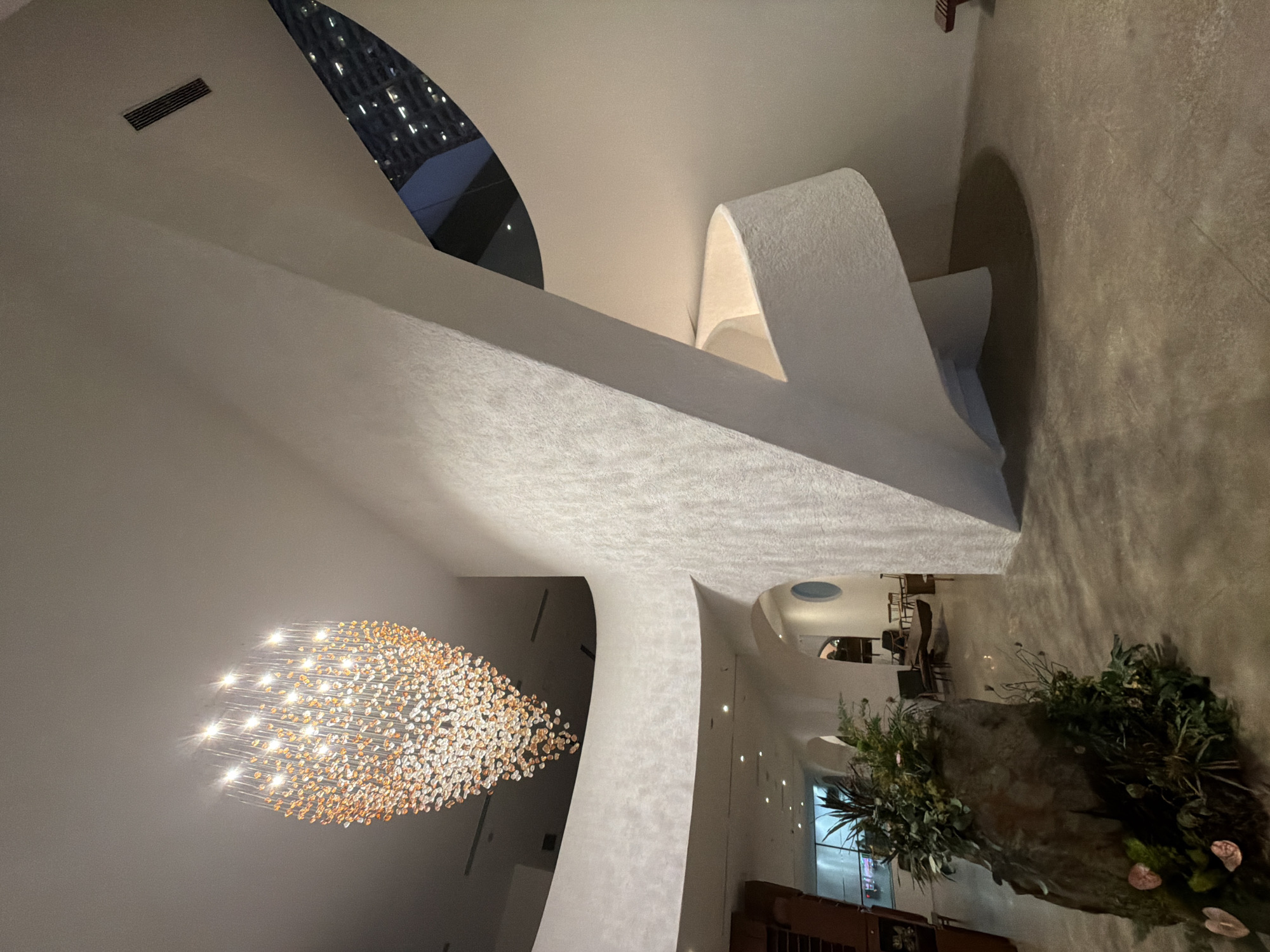 The image shows the interior of a modern building. A large, round chandelier hangs from the ceiling. The walls are white and curved. A window with a view of a building is visible. The floor is stone and a large floral arrangement sits on it. Furniture is visible in the background. The location is likely in Nha Trang Ward, Vietnam.
