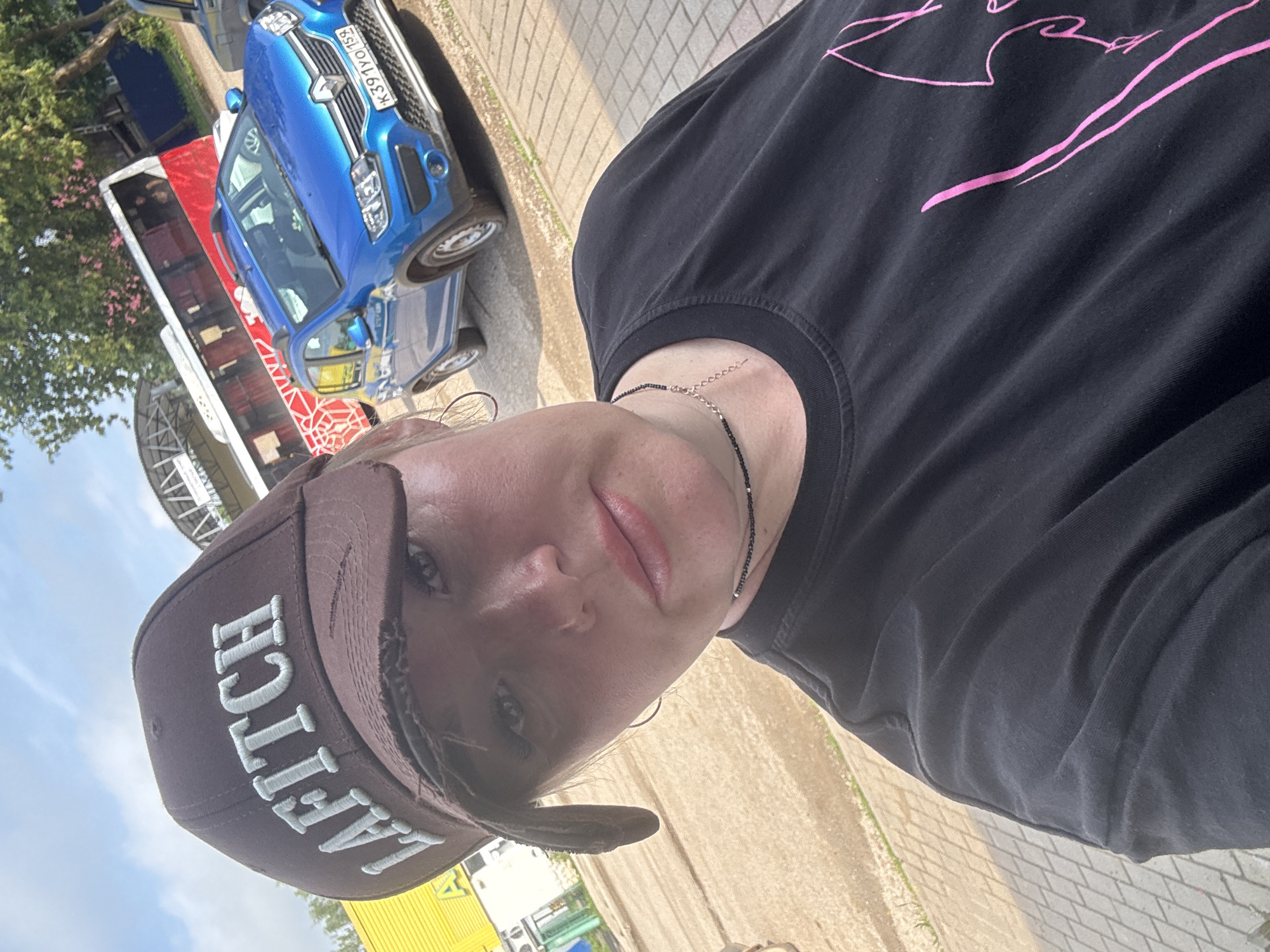 A woman is taking a selfie outdoors in Chastyye, Russia. She is wearing a brown baseball cap with the word "LAFITICH" embroidered on it in white letters. She also has on a black t-shirt and a thin black necklace. In the background, a blue Renault Sandero car is parked, and a red and white bus is visible. The woman is standing on a paved area with a textured surface, possibly a sidewalk or parking lot. The sky is blue with some clouds, and trees can be seen in the distance. The overall scene suggests a casual, everyday setting.