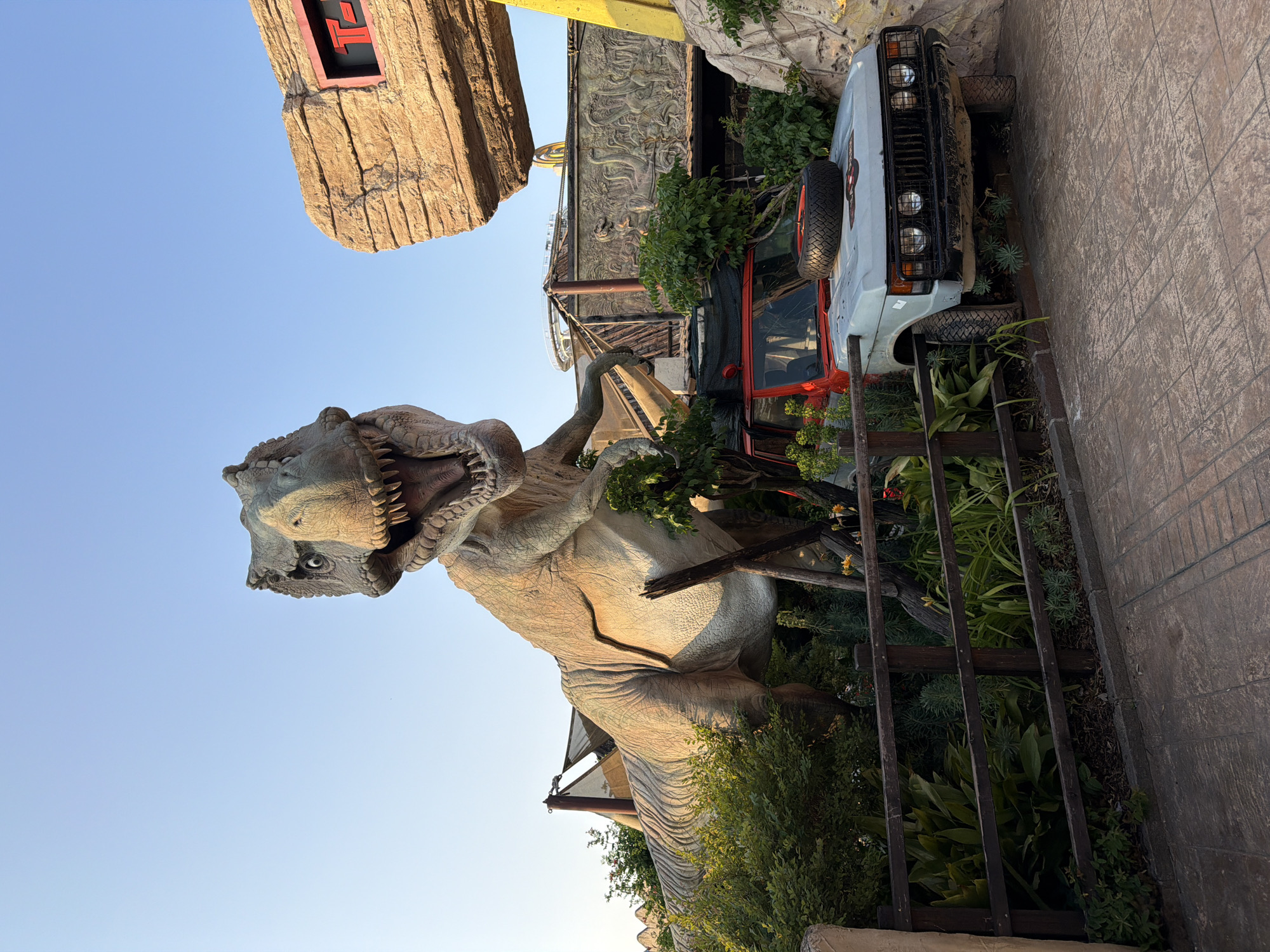 A life-sized replica of a Tyrannosaurus Rex dinosaur stands imposingly in front of a pale blue sky, its mouth open in a roar. The dinosaur is positioned behind a wooden fence and lush green foliage, with a weathered, light-colored off-road vehicle parked to its right. The vehicle has a red roof and a spare tire mounted on its hood. In the background, structures resembling rock formations and themed park elements are visible, including a sign with a red "T" and part of a roller coaster track. The scene appears to be an outdoor attraction, likely in a theme park, given the stylized scenery and the presence of dinosaur models. The overall impression is one of a Jurassic Park-themed exhibit. This exhibit is located in Gardaland, Castelnuovo del Garda, Italy.