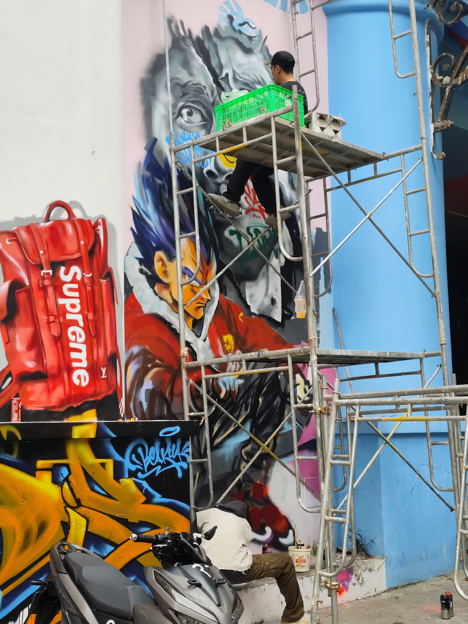 The image captures a street art mural in progress, likely in Da Lat, Vietnam. The mural features a stylized rendering of Vegeta from Dragon Ball, alongside a Supreme-branded Louis Vuitton backpack and other graffiti elements. Two individuals are actively working on the mural, using scaffolding for access. One person, wearing a black shirt and cap, stands on the scaffolding with a green basket. Another person wearing a white shirt is crouched near the bottom of the mural. A motorcycle is parked in front of the wall, and cans of spray paint are scattered around. The background includes a light blue pillar and a white wall. The scaffolding is standard metal construction.