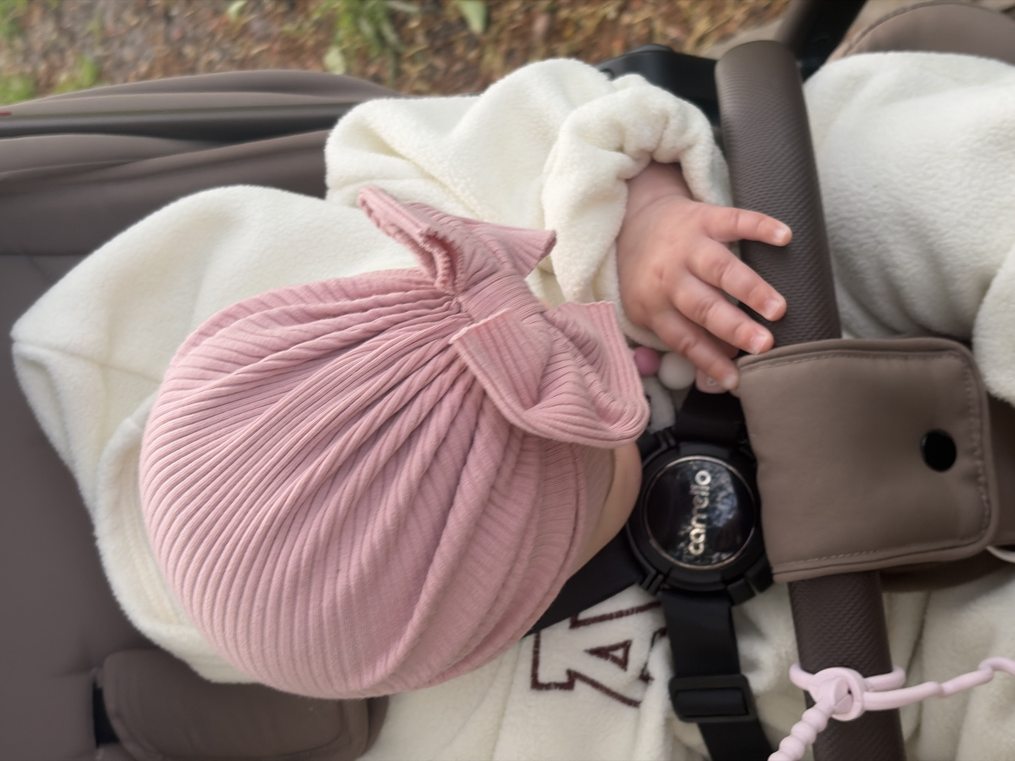 A top-down, medium shot depicts an infant seated in a stroller. The infant is wearing a white, textured jacket and a pink, ribbed knit hat that resembles a tied bundle. The baby's hand rests on the stroller's padded bar, near a black wristwatch with the word "Carrello" visible on the watch face. A brown pouch is affixed to the stroller's bar. A portion of the stroller's brown seat and frame are visible. Pink stroller clips are knotted around the bar. The location cannot be determined from the image.