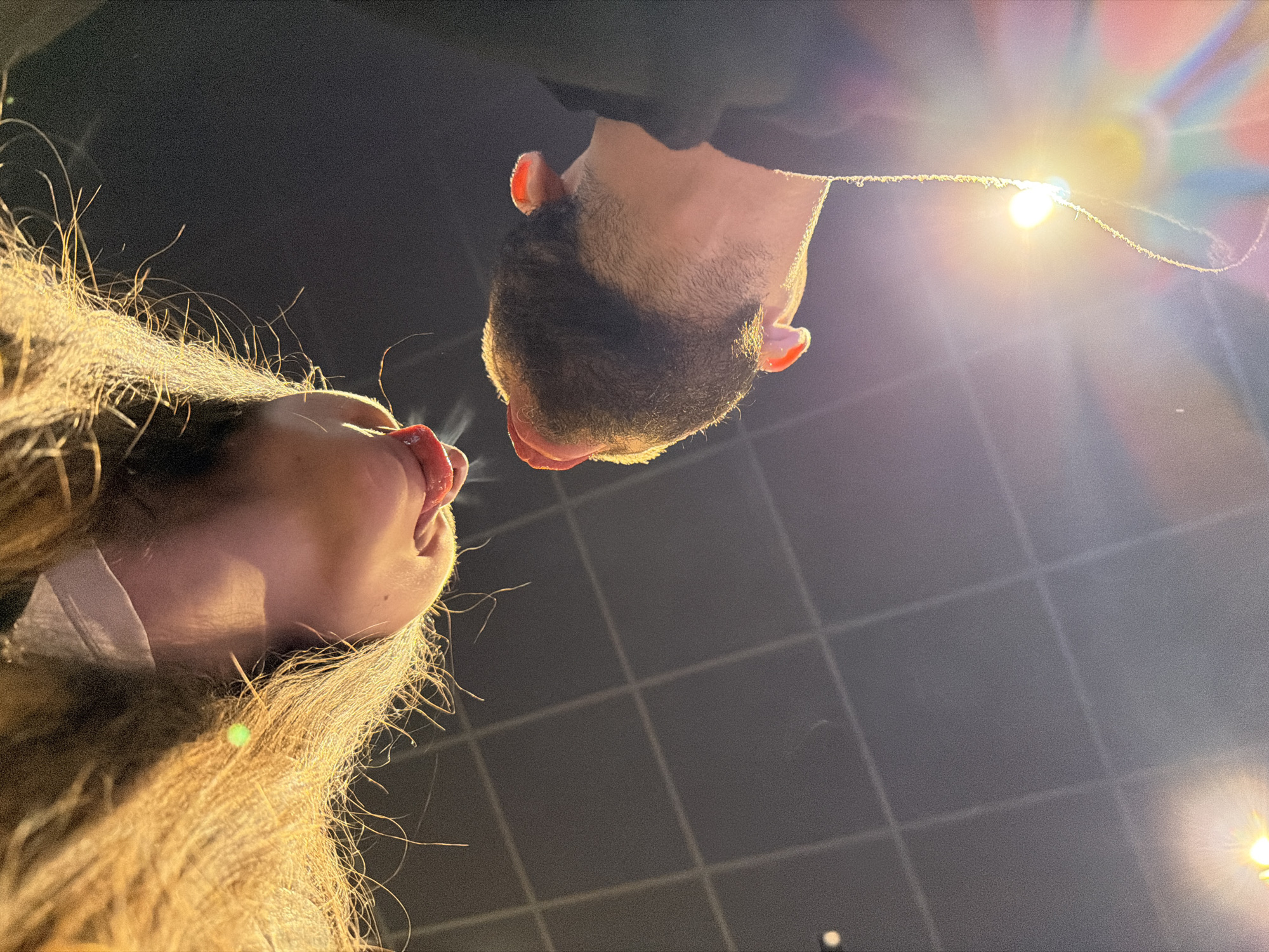 Two individuals, a man and a woman, are positioned close to each other, their heads oriented towards the center of the frame, tongues out. The man has dark hair and a beard. The woman has long brown hair. A bright light source is visible above them. The background is a dark tiled ceiling. There is no definitive evidence to conclude that this image was taken in the city of Pernik, Bulgaria.