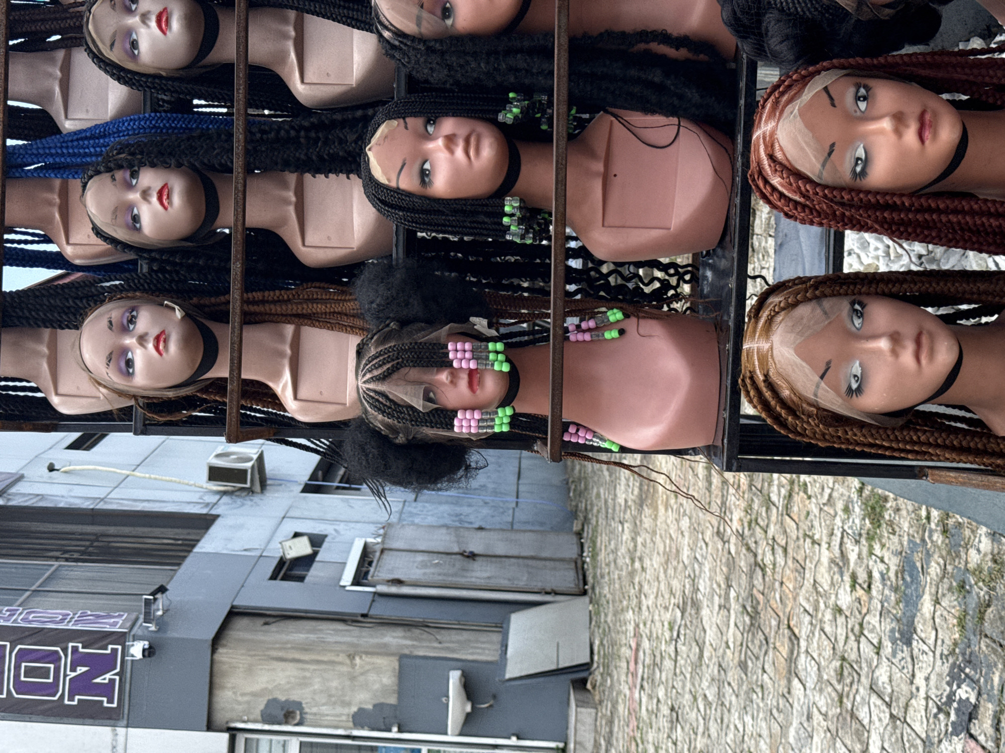 The image depicts a street scene in Ado, Nigeria, featuring a metal rack displaying mannequin heads adorned with various braided wigs. The mannequin heads have painted faces and black chokers. The wigs are in shades of black, brown, and blue, with some incorporating colorful beads. The background consists of a multi-story building with air conditioning units and a business sign with the text "ION". The ground is paved with interlocking stones. No people are visible in the scene.