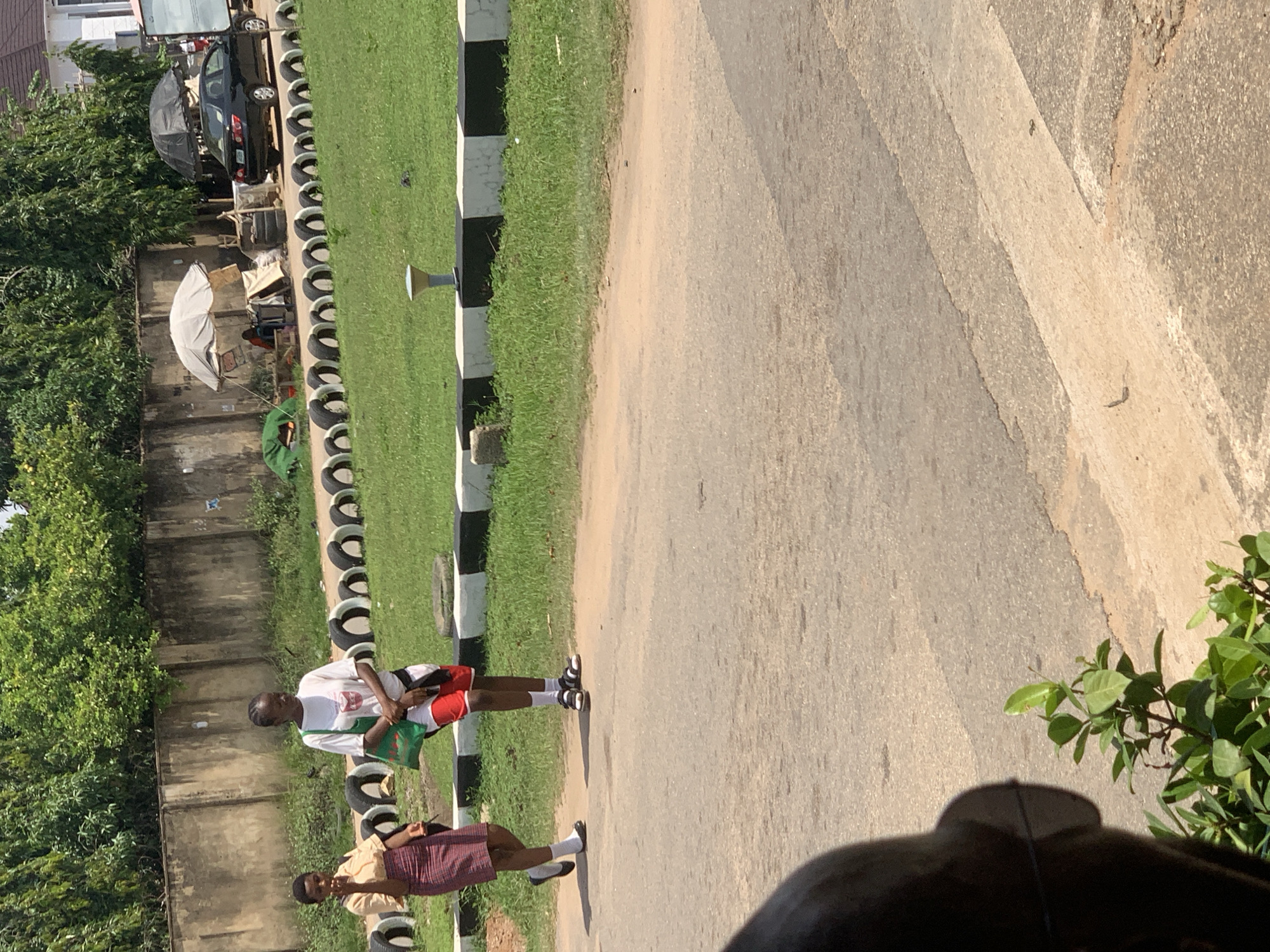 In Abuja, Nigeria, two young women are walking on a paved road. The woman on the left wears a cream-colored short-sleeved shirt and a red and white plaid skirt. She carries a black bag. The woman on the right wears a white t-shirt with a graphic print and red shorts. She has a green bag slung over her shoulder. They are walking side-by-side.
Behind them, a low concrete wall runs parallel to the road. Beyond the wall, there is a grassy area with a line of black and white-banded curbs. Further back, a black car is parked under a black umbrella. Next to the car is a larger, light-colored umbrella covering a small market stall with goods displayed. Trees and bushes form a dense background, with a building visible in the far right. The ground in the foreground is paved asphalt.