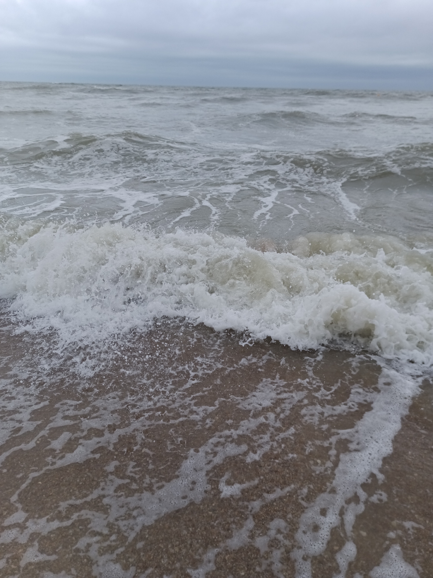 A choppy, grey ocean meets a sandy shore under an overcast sky. Waves crash and foam as they recede, leaving a wet, textured sand with a sparse scattering of foam. The horizon is indistinct, blurred by the hazy atmosphere and the rough sea. The scene evokes a sense of a blustery day in Houlgate, France, likely near the Normandy coast.