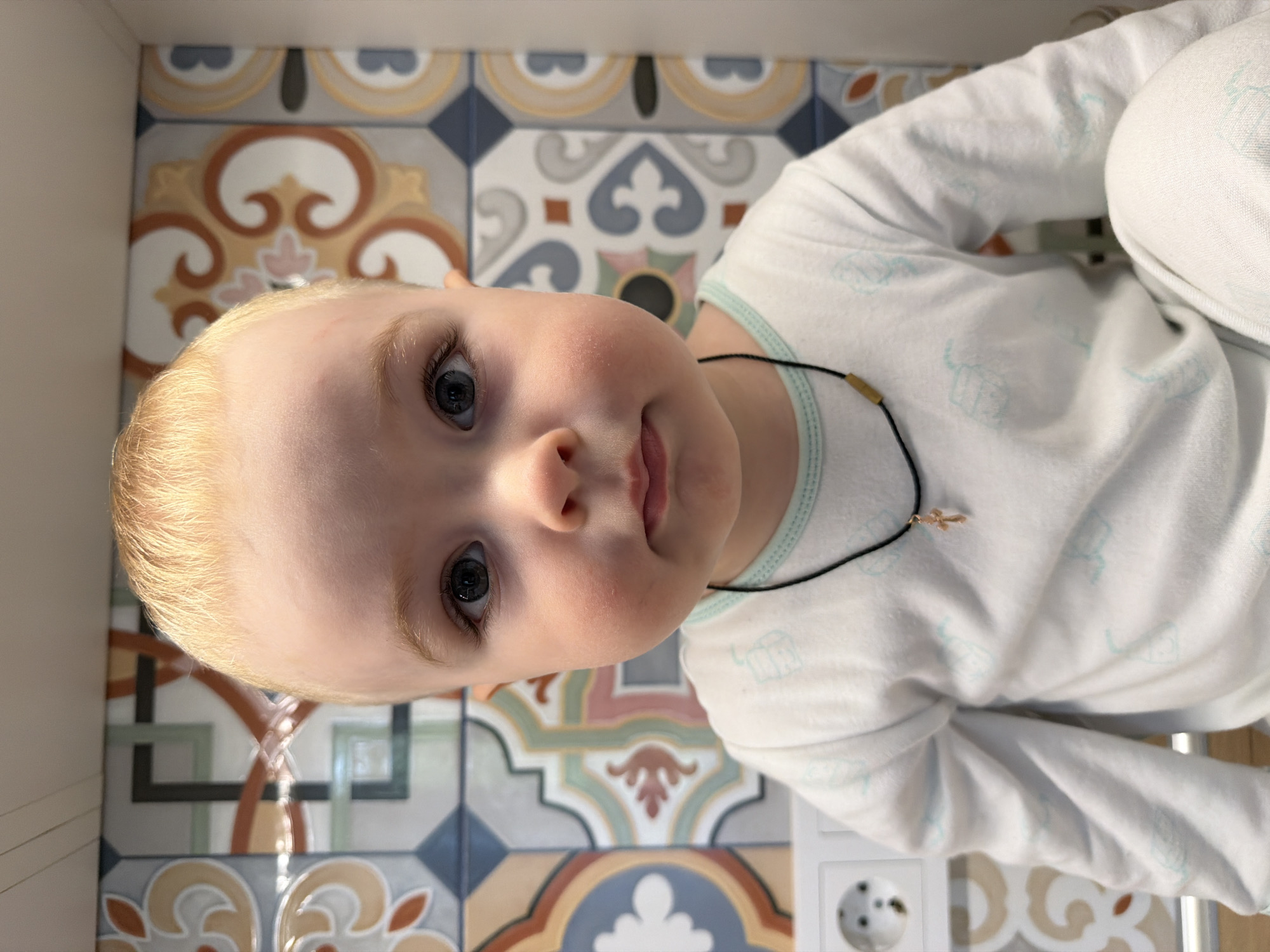 A baby with blond hair and blue eyes is positioned in front of a tiled surface featuring a repeating pattern of geometric shapes and floral motifs in muted colors. The baby is wearing a white long-sleeved shirt with light blue patterns and a thin black necklace with a small gold cross pendant. A white electrical outlet is visible in the lower right corner of the frame. The location is not evident from the image content alone to determine if it is in Votkinsk, Russia.