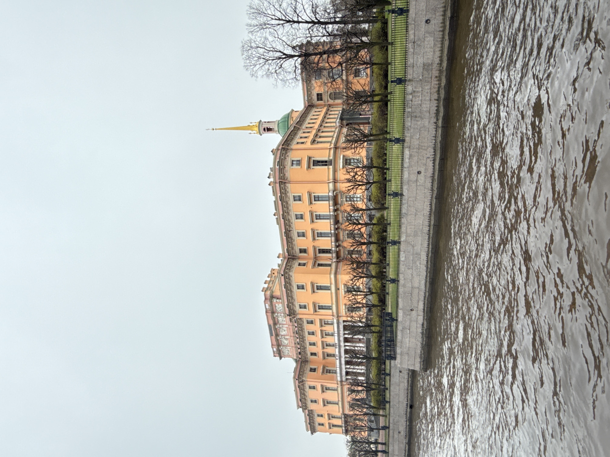 The image captures a view of the Mikhailovsky Castle in Saint Petersburg, Russia, along the Moika River. The castle's facade is pale orange with architectural details, including numerous windows and decorative elements. A green dome topped with a golden spire is visible, indicating the location of the church or chapel within the castle complex. Bare trees line the embankment in front of the castle, separating it from the water's edge. A metal fence runs along the embankment, providing a barrier between the pathway and the river. The water of the Moika River is murky and reflects the overcast sky. The sky itself is a uniform pale gray, suggesting a cloudy day. The perspective is from the river, likely taken from a boat or from the opposite bank. There are no visible people or activities in the scene beyond the static architectural view.
