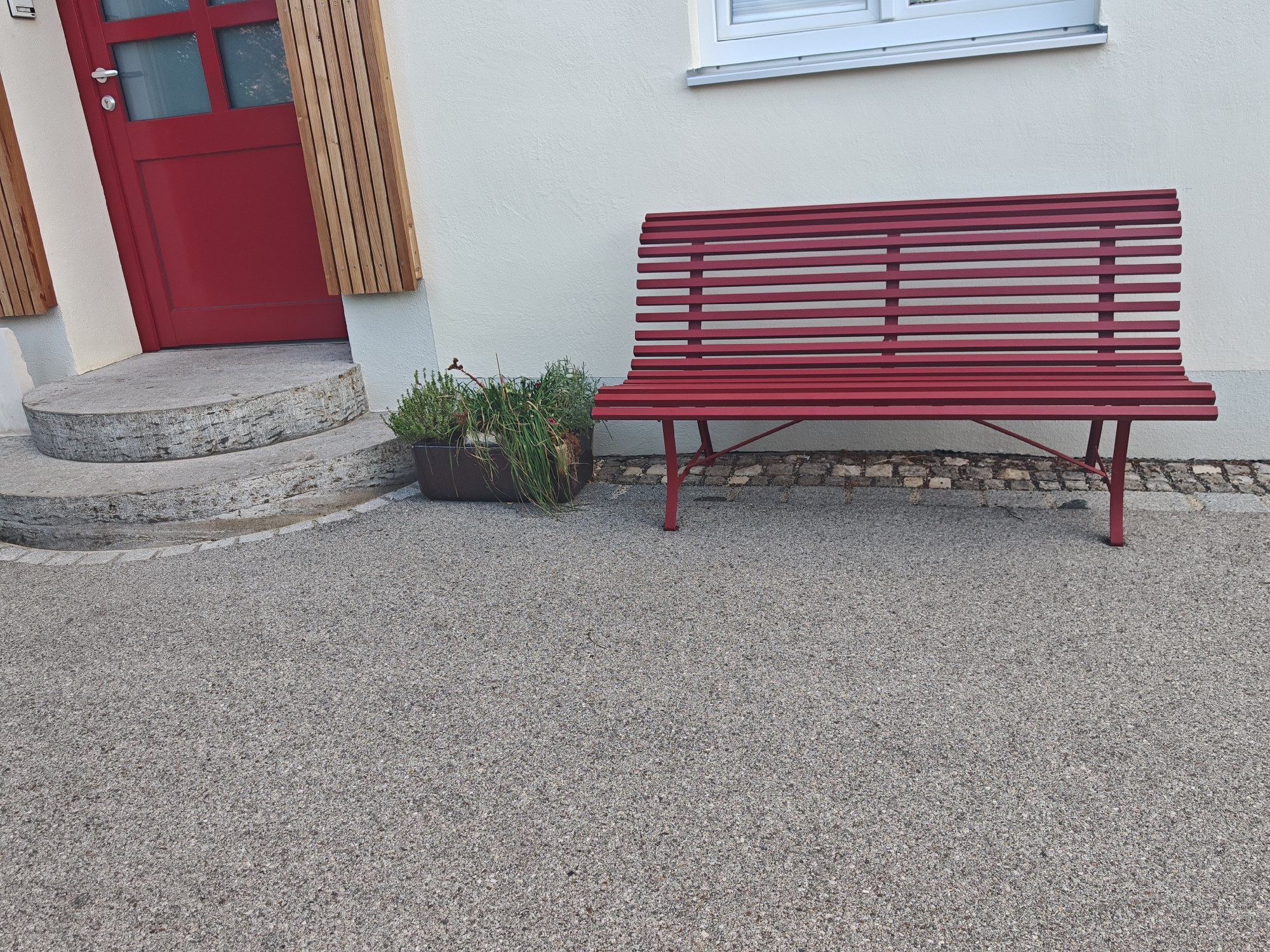 The image shows an exterior view of a building in Obergünzburg, Germany. A bright red door with glass panes and wooden accents is visible on the left side, with a curved stone step leading up to it. To the right of the door, a dark planter contains green foliage. A red slatted bench is positioned in front of a cream-colored wall, under a horizontal window. The ground is covered in a speckled grey gravel-like material, with cobblestones visible near the bench. The scene is static and appears to be a quiet, residential or commercial exterior.
