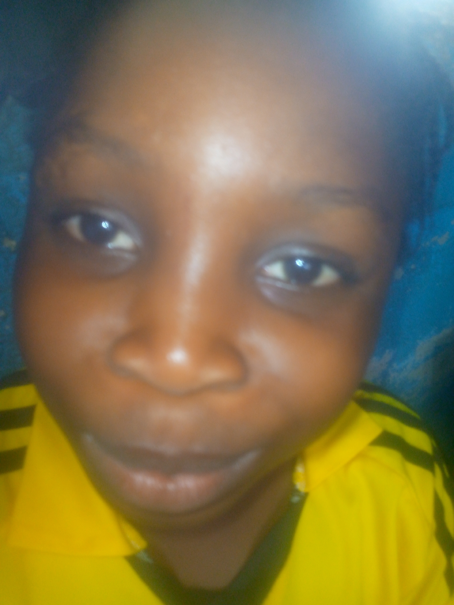 A close-up photograph shows a child with dark skin wearing a yellow shirt with black stripes. The child is positioned indoors, likely in Ikorodu, Nigeria. The background is a blue wall. The child is looking directly at the camera.