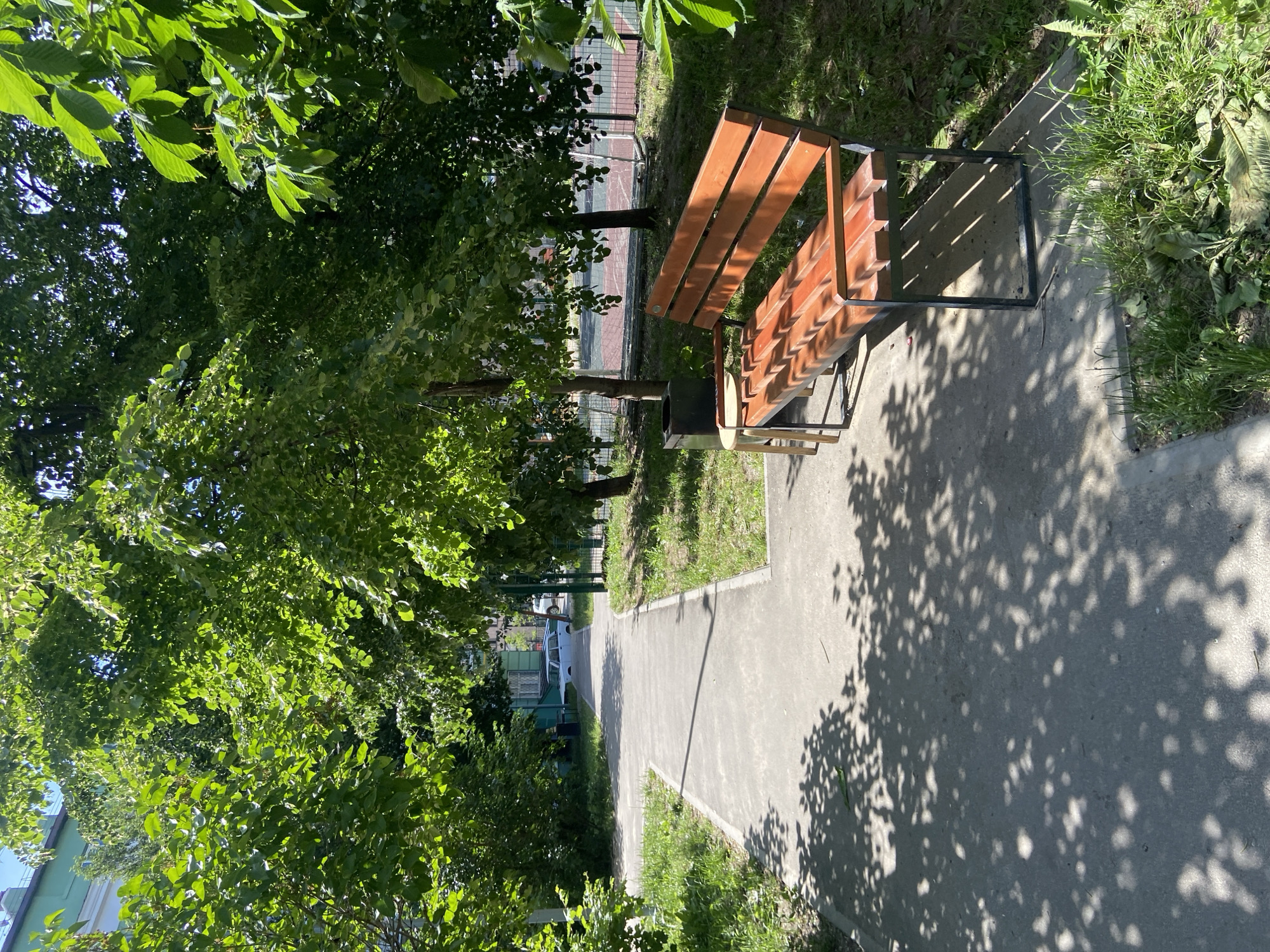 This image depicts an outdoor pathway scene, likely within a park or residential area in Kazan, Russia, under bright sunlight. A concrete or paved walkway, partially shaded by numerous trees, extends from the foreground and curves slightly to the left into the background. On the right side of the pathway in the mid-ground, a wooden slatted bench with a dark metal frame is situated. Adjacent to the bench's left side is a small, light-colored stool or table, and behind it, a dark square trash can.
Dense green foliage from deciduous trees frames the scene, casting distinct, dappled shadows across the pathway and ground. Patches of green grass and ground cover are visible alongside the path. In the background, to the left, a light-colored sedan (resembling an older model, possibly a Lada) is parked, with a building featuring a green facade visible behind it. To the right in the background, beyond a chain-link fence and trees, a sports court with red and green surfacing is visible, including elements suggesting a goalpost or net. A small portion of clear blue sky is discernible through the tree canopy. No individuals are present in the scene.