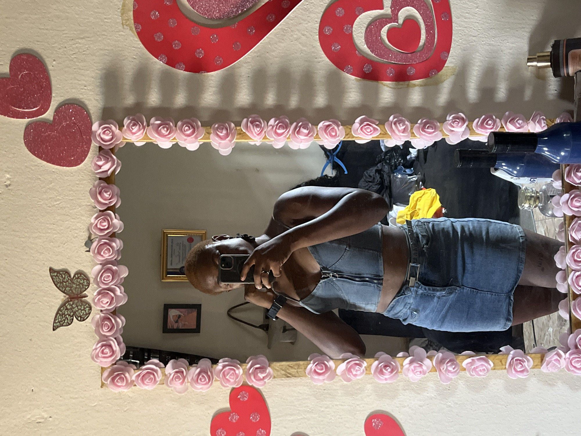The image captures a mirror selfie taken by a woman in an indoor setting, possibly in Jalingo, Nigeria. The photograph is oriented sideways, with the subject appearing horizontal.
The woman, with dark skin and short hair, is holding a smartphone to capture her reflection. She is wearing a sleeveless denim-like crop top with a front zipper and a matching denim mini skirt. A watch is visible on her left wrist.
The mirror she is using is elaborately decorated. Its frame is adorned with numerous light pink rose embellishments. The wall around the mirror, which is a light, neutral color, features additional decorative elements. Above and to the left of the mirror, there are several red heart cutouts, some with a glittery texture. A metallic, intricately designed butterfly decoration is placed on the wall to the left of the mirror.
Reflected in the mirror's surface, behind the woman, are various items. These include a gold-framed picture, a smaller dark-framed picture, several bottles (some blue), and what appears to be black plastic bags and a yellow piece of fabric. The overall scene suggests a personal and decorated space. No discernible text is visible in the image.