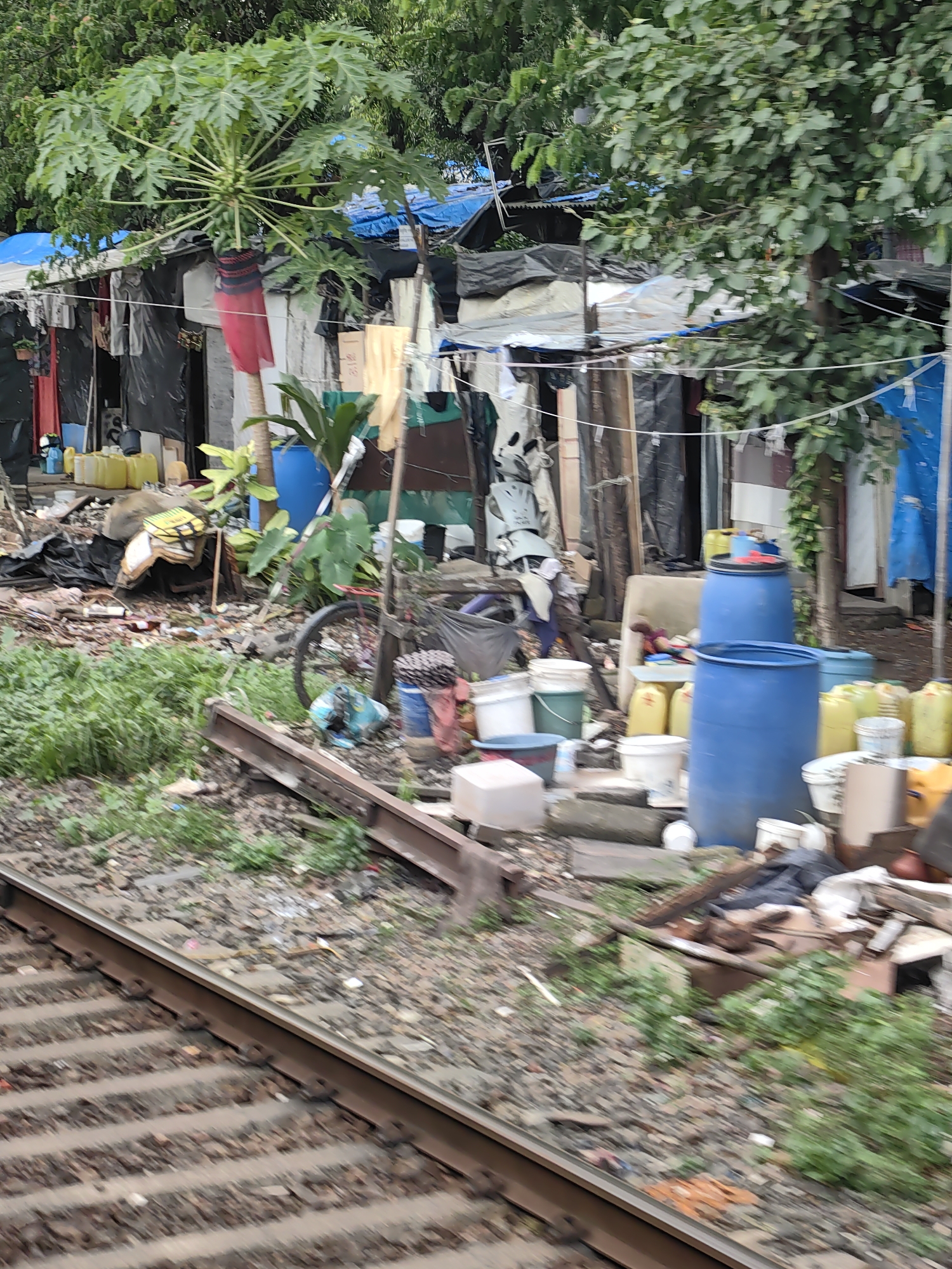 The image depicts a slum settlement adjacent to railway tracks in Mumbai, India. The settlement consists of makeshift shelters constructed from tarpaulins, plastic sheeting, and various salvaged materials, creating a dense and cluttered environment. Laundry hangs on lines strung between structures, indicating daily life. Various objects are strewn about, including buckets, containers, bags of materials, and discarded items, contributing to the scene's disarray. A bicycle with a partially visible wheel is leaned against a structure. The foreground shows the railway tracks, indicating the proximity of transportation infrastructure to the settlement. The overall impression is one of a densely populated, informal living space situated in an urban environment.