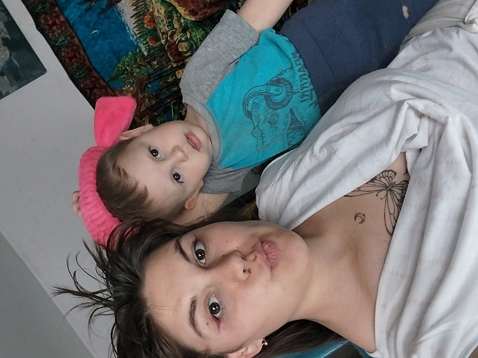 A selfie shows a woman and a young child. The woman is in the foreground, looking at the camera with her lips pursed. She has dark hair, brown eyes, and is wearing a white shirt. A butterfly tattoo is visible on her chest. To the woman's right and slightly behind her is a young child with brown hair and brown eyes, wearing a blue and gray shirt. The child is looking towards the camera. A pink fuzzy headband is on the child's head. They appear to be indoors, with a patterned blanket in the background. The location is Unknown, Unknown.