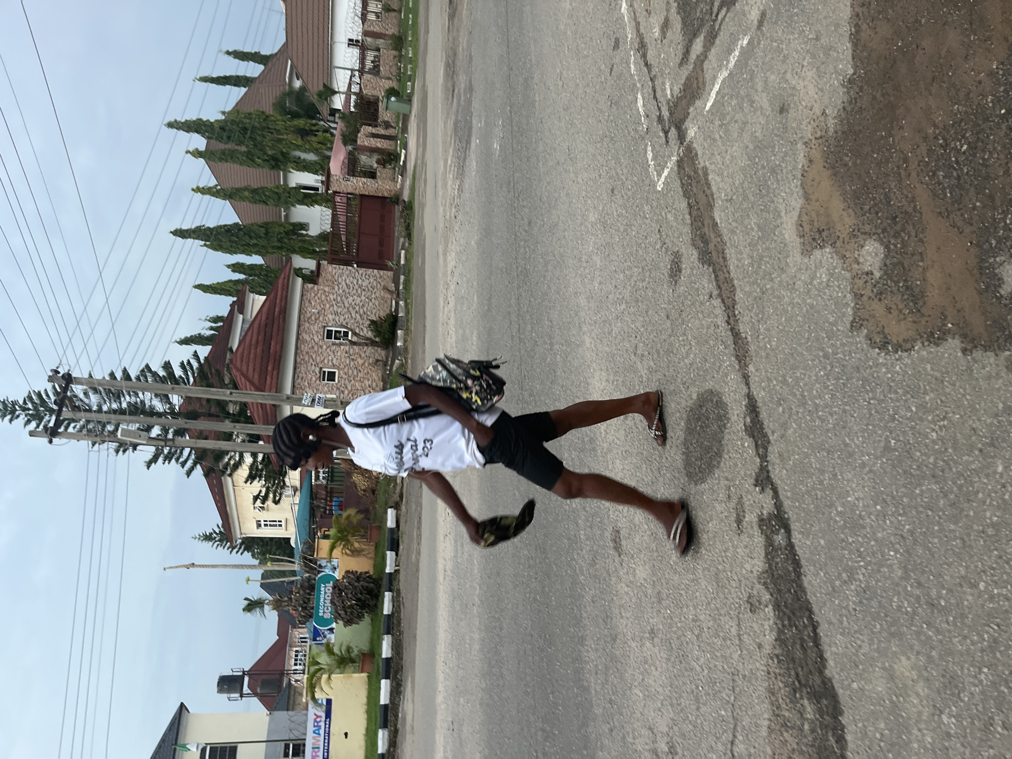 A medium shot captures a young Black woman walking from left to right across a paved road in Abuja, Nigeria. She is wearing a white t-shirt with black text, black shorts, and flip-flops. She carries a black patterned backpack on her left shoulder and holds a dark-colored item, possibly a shoe or a hat, in her right hand. Her braided hair is visible. The background features a variety of residential buildings with tiled roofs, some with fences and gates, as well as utility poles with overhead wires and evergreen trees. A sign for "PRIMARY SCHOOL INTERNATIONAL" is visible on the left. The road shows some wear and tear, with patches of discoloration and a puddle of water. The sky is overcast.