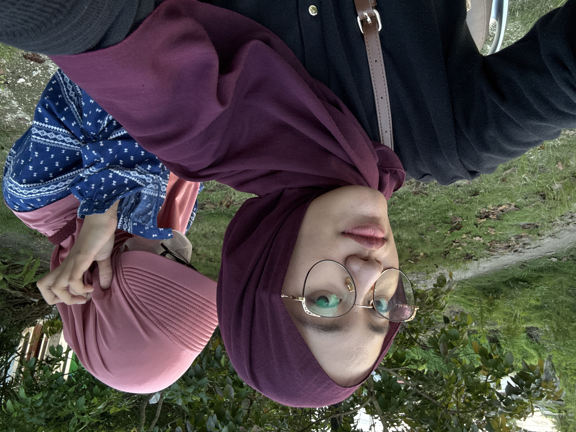 Two women are outdoors, near vegetation, in Gadingrejo, Indonesia. One woman wears a purple hijab and gold-rimmed glasses. The other wears a pink hijab and a blue patterned blouse. One woman's arm and a black shirt with a brown strap and gold buttons are visible. Grass and a path are in the background.