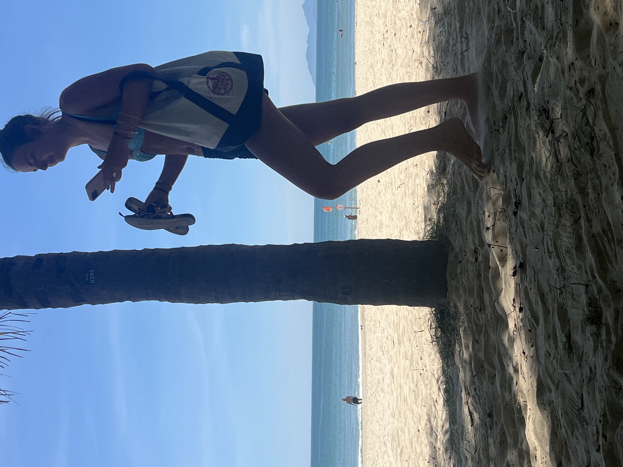 A young woman, wearing a teal bikini top and dark shorts, stands on a sandy beach in Da Nang, Vietnam, near a palm tree. She is looking at her phone, which she holds in her right hand. In her left hand, she carries a pair of flip-flops. A beige tote bag with dark trim hangs from her left shoulder. The beach is bathed in sunlight, casting shadows from the palm tree onto the sand. The ocean stretches out behind her, with a calm blue horizon. In the distance, a man is visible wading in the water. The sky is clear and blue. The image is taken from a low angle, looking up towards the woman and the sky.
