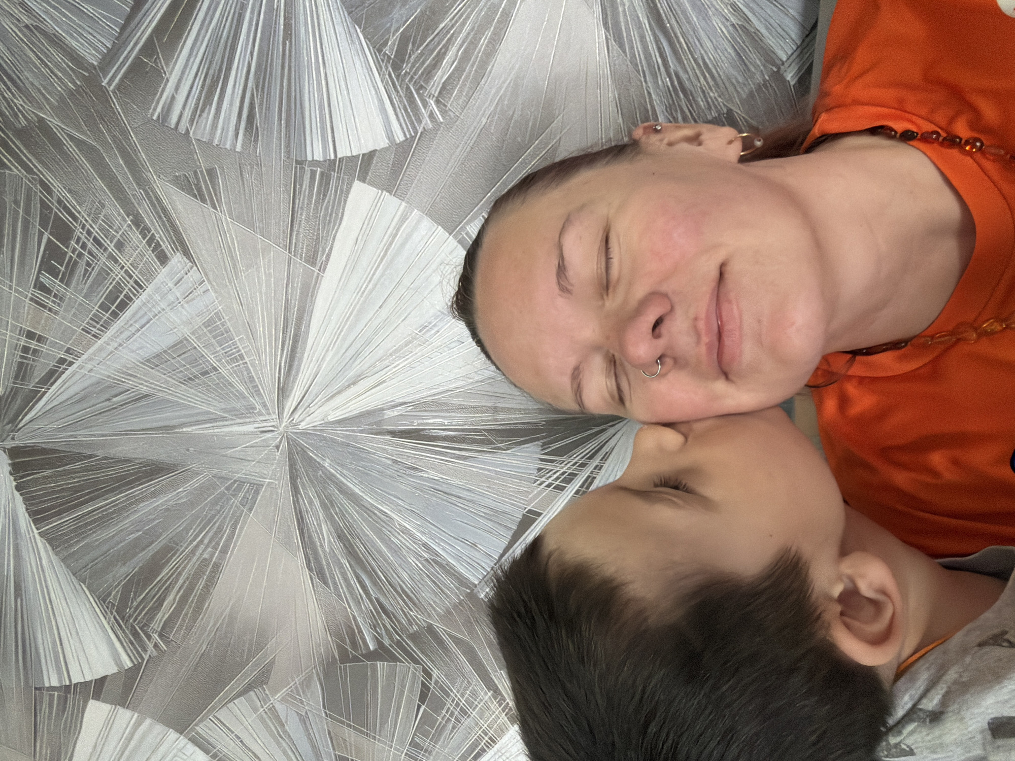 A photograph shows a woman and a child lying down with their eyes closed, apparently resting. The woman is wearing an orange shirt and a necklace with amber-colored beads. She has multiple piercings in her ear and a ring in her nose. The child, who seems to be a boy, is wearing a gray shirt. They are lying on a surface with a geometric, stylized sunburst or starburst pattern. The setting is identified as Chaikovsky, Russia.