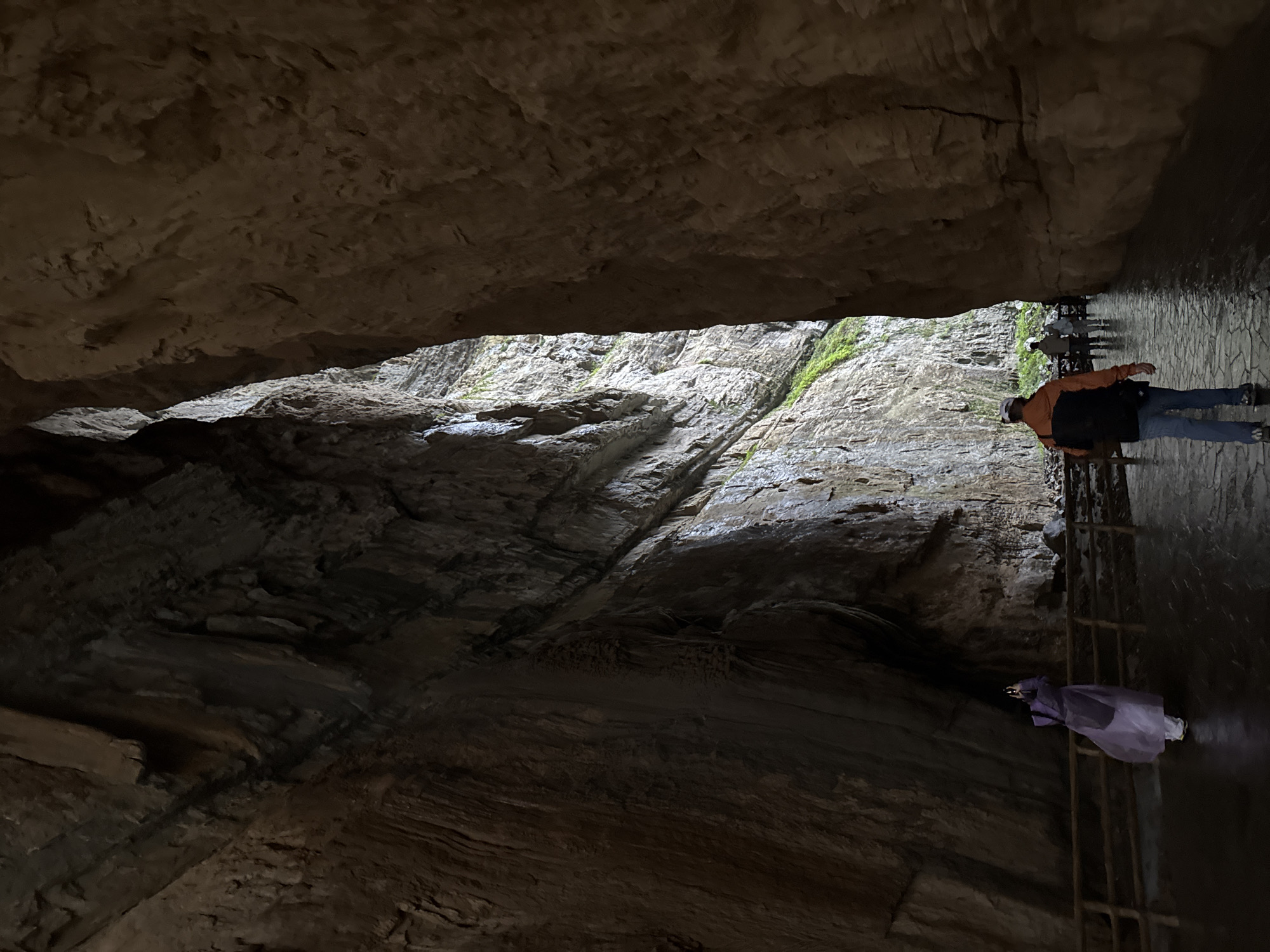 The image depicts a scene inside a cave at the Wulong Karst geological park in Wulong District, Chongqing, China. Two individuals are visible along a narrow stone pathway. One person is wearing an orange jacket and walking, while the other is wearing a purple poncho and appears to be taking a photo. The pathway is adjacent to a rocky cave wall with a stream running down. The roof of the cave is close to the pathway, creating a low ceiling.