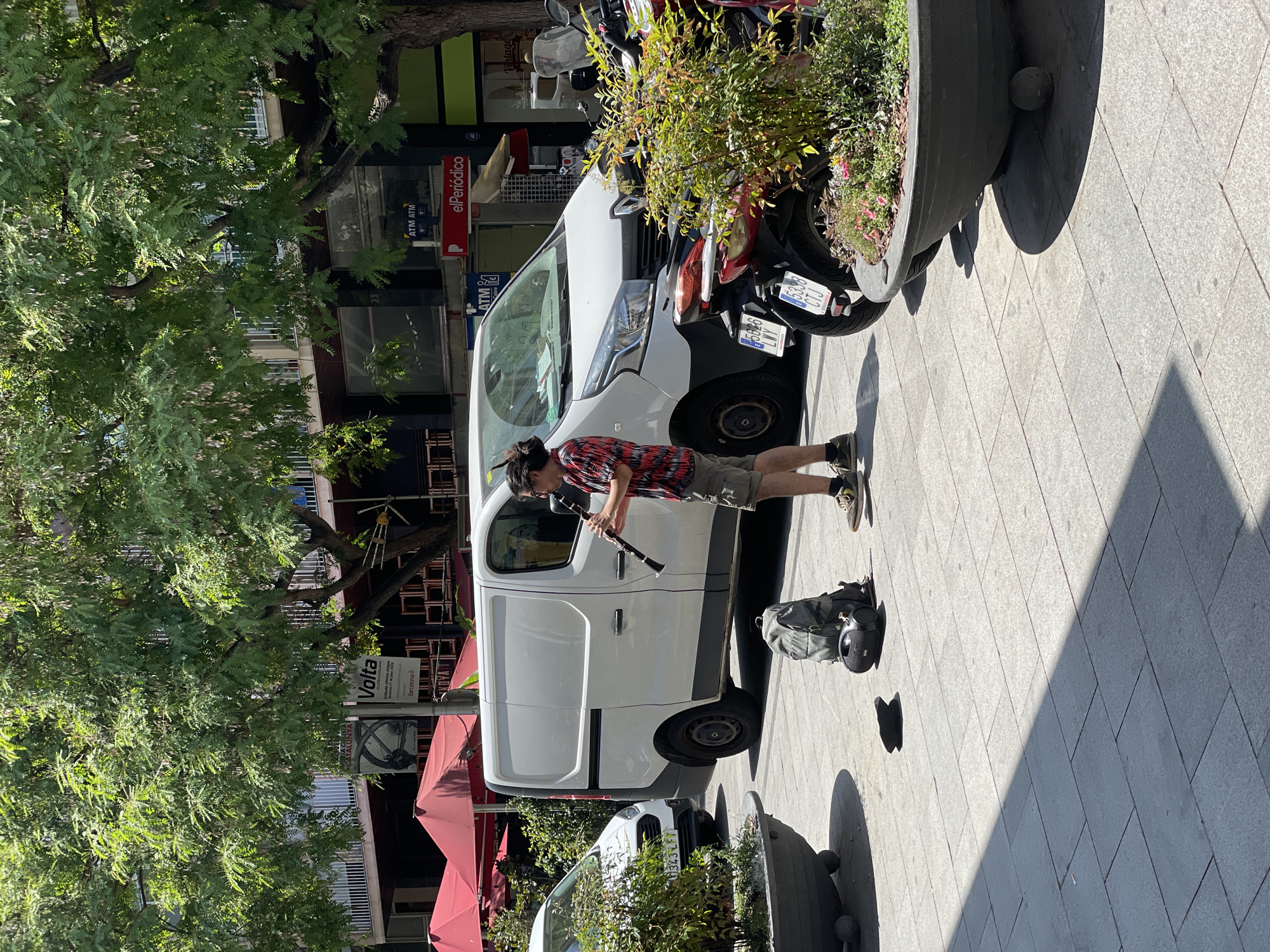 A person is playing a clarinet on a sun-drenched, tiled sidewalk in Barcelona, Spain. The musician, wearing a red and white checkered shirt and grey shorts, stands next to a white van and a motorcycle. A backpack and a small boombox are on the ground beside them. In the background, trees provide shade over buildings with shops and awnings. A sign for "Volta" is visible on a building. The scene captures a moment of everyday urban life and street performance.