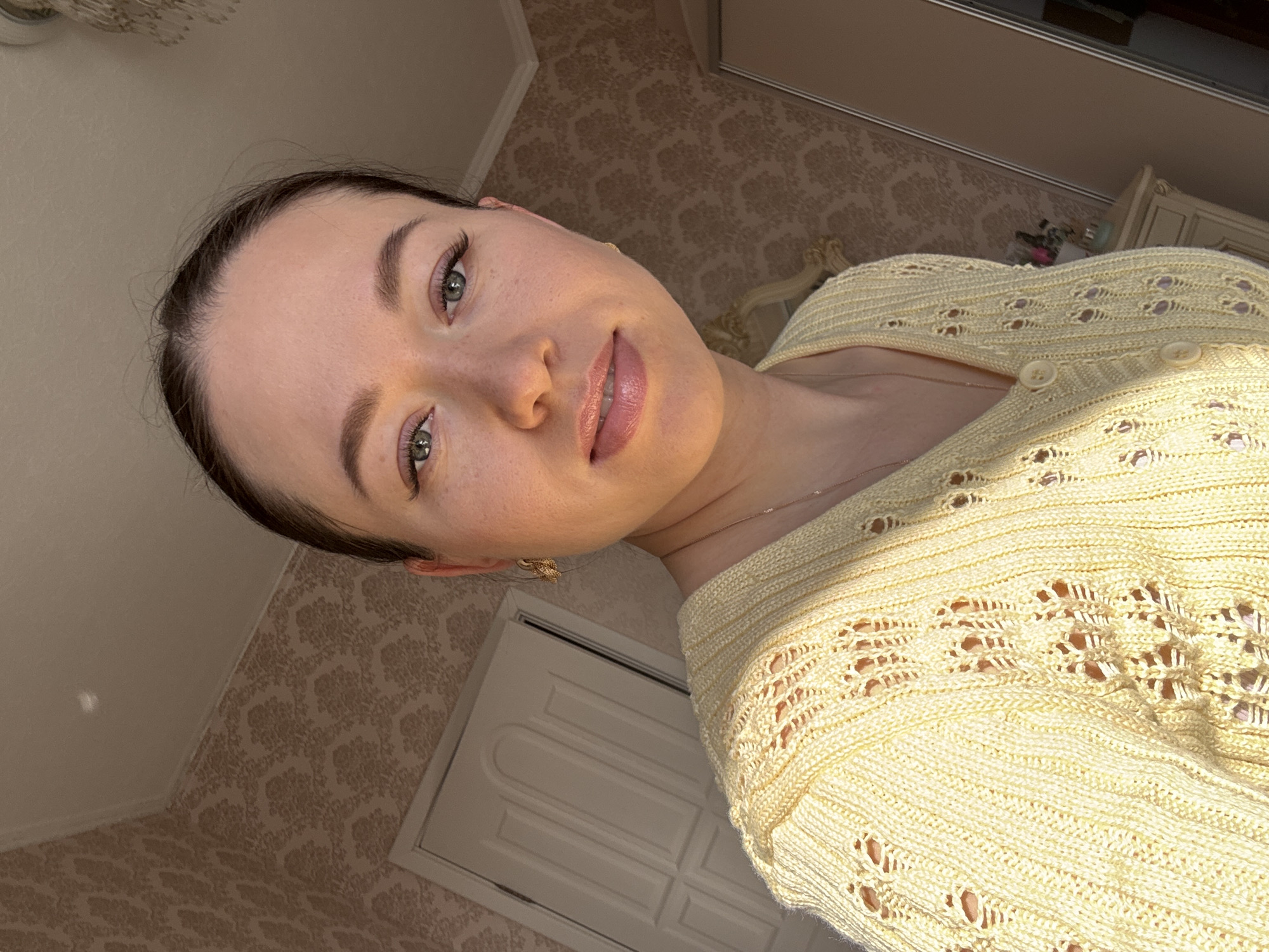 A woman with fair skin and dark hair pulled back poses indoors. She wears a light yellow knit sweater with decorative cutouts. Her eyes are light-colored, and she has light pink lipstick. A small, gold-colored earring is visible in her left ear. The background features patterned wallpaper and a white door. A partially visible dresser with decorative elements is also present. The image appears to be taken at an angle, looking down slightly at the subject. There is no indication the image was taken in Ferma, Russia.