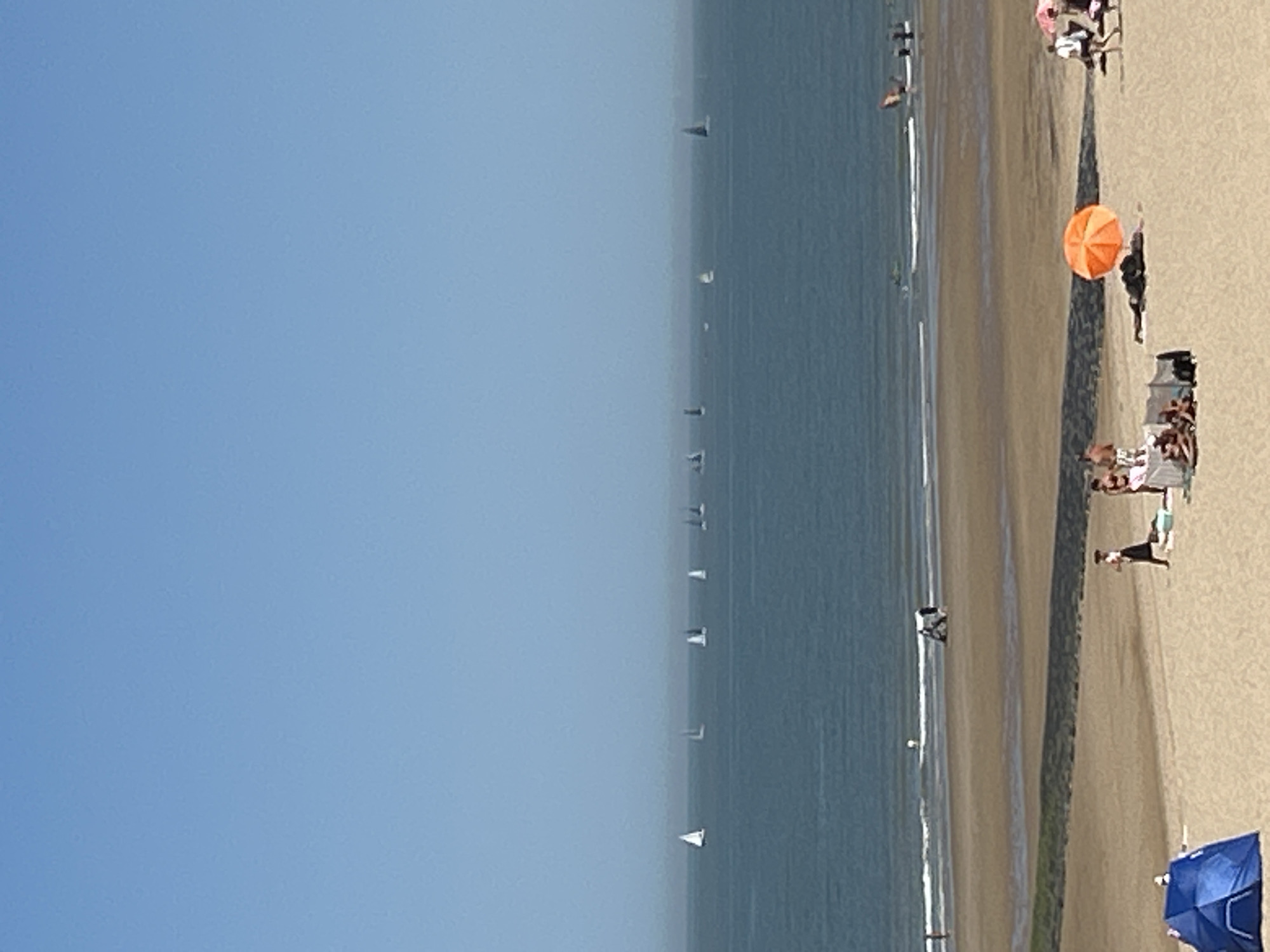 This image depicts a sunny day at the beach in Ostend, Belgium. The scene is divided into three main sections: the sky, the sea, and the beach.
The sky is a clear, pale blue, taking up the upper half of the image. The sea is a deep blue-green, with a hazy horizon. Numerous sailboats with white sails are scattered across the water, suggesting a sailing regatta or leisurely sailing activity. Some boats are closer to the shore, while others are further out.
The beach in the foreground is sandy and appears to be quite wide. There are several groups of people present. On the beach, a blue tent is set up on the left side. Further to the right, a group of people are gathered around an orange umbrella, with some lying down and others sitting. Another individual is walking away from the camera. In the middle ground, near the water's edge, a few people are walking and playing. Some figures are visible in the water, and there appears to be some form of water sports occurring, possibly windsurfing or kiteboarding, indicated by a sail-like object in the distance. The tide is out, exposing wet sand and shallow pools. A dark, textured line, possibly a breakwater or retaining wall, separates the drier sand from the wetter sand closer to the sea. The overall atmosphere is one of a relaxed summer day at the coast.