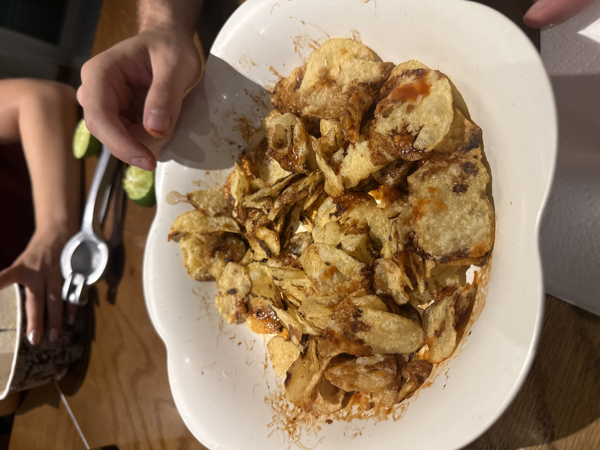The image captures an overhead view of a casual indoor setting, likely a dining or snack occasion. The central subject is a white, wavy-edged bowl filled with numerous potato chips, which appear to be covered in a dark, glistening sauce or seasoning, and some also have distinctive reddish spots of what seems to be hot sauce or ketchup. The chips vary in shape and browning, suggesting they might be homemade or specialty fried potatoes.
Two individuals are partially visible. On the left, a person's bare arm and hand (likely male, indicated by arm hair) is reaching into the bowl, poised to grab a chip. To the left and slightly below this person's hand, another person's hand (likely female, indicated by light-colored nail polish) is visible, holding a silver metal citrus squeezer. Next to the squeezer and the hand are at least two sliced green limes, suggesting fresh citrus is being prepared, possibly for the chips or a drink.
The items are arranged on a wooden table surface. A light-colored disposable cup or container is partially visible in the lower-left corner, and a white paper towel or napkin can be seen on the far right edge of the frame. The lighting appears to be indoor artificial light, creating some shadows. No visible text is present in the image. The overall scene conveys a relaxed, informal atmosphere focused on sharing food.