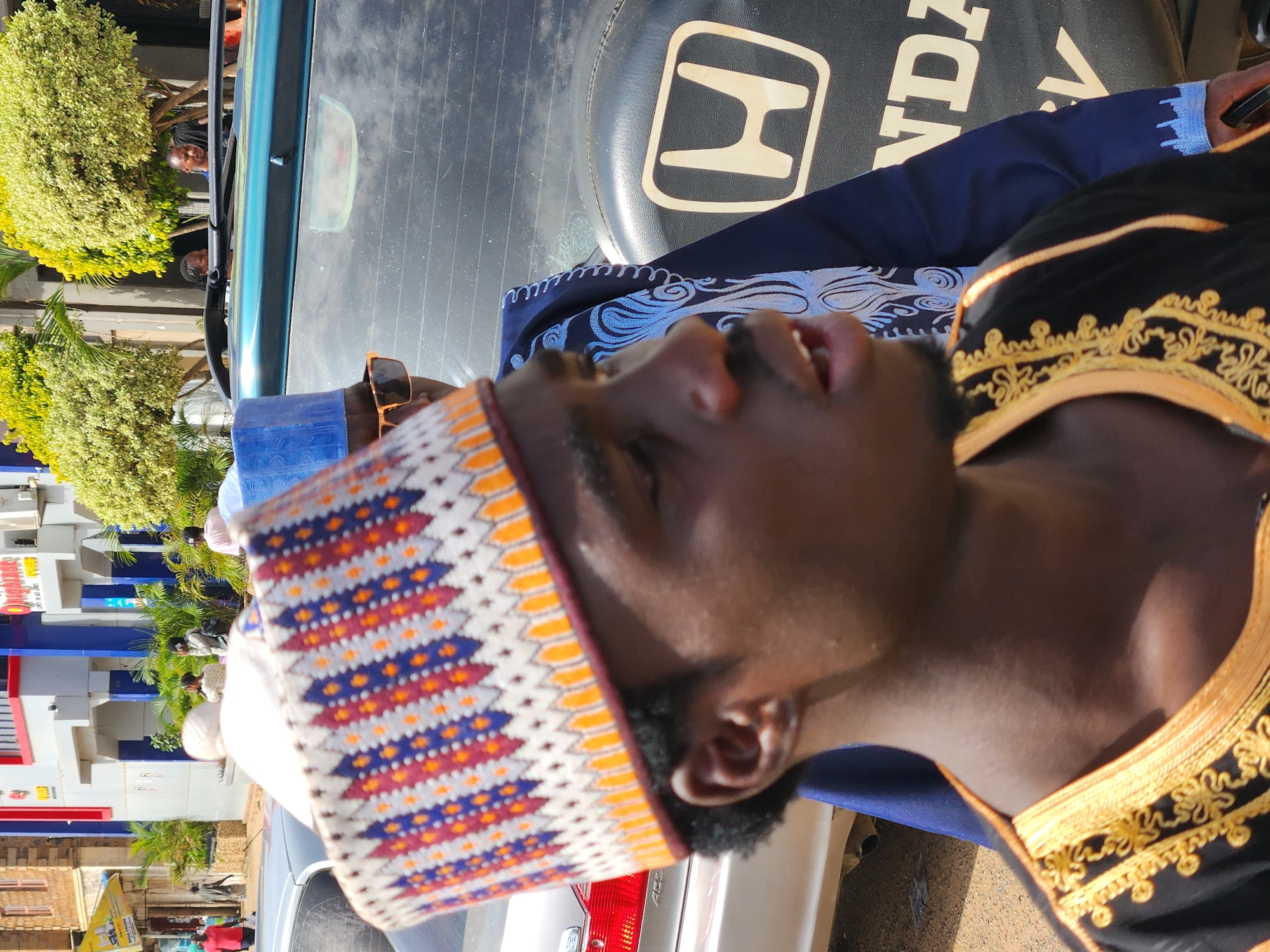 A man in a patterned hat and embroidered clothing stands in front of a parked vehicle in an urban environment in Unknown, Unknown. The man is wearing a traditional cap with orange, white, and blue geometric patterns. He also wears clothing with gold-colored embroidery on a black background. The vehicle behind him has a spare tire cover featuring the Honda logo. Behind the car, there is a building with blue and white walls and a sign that partially reads "Supermarket." People are standing around the corner of the building near some greenery.