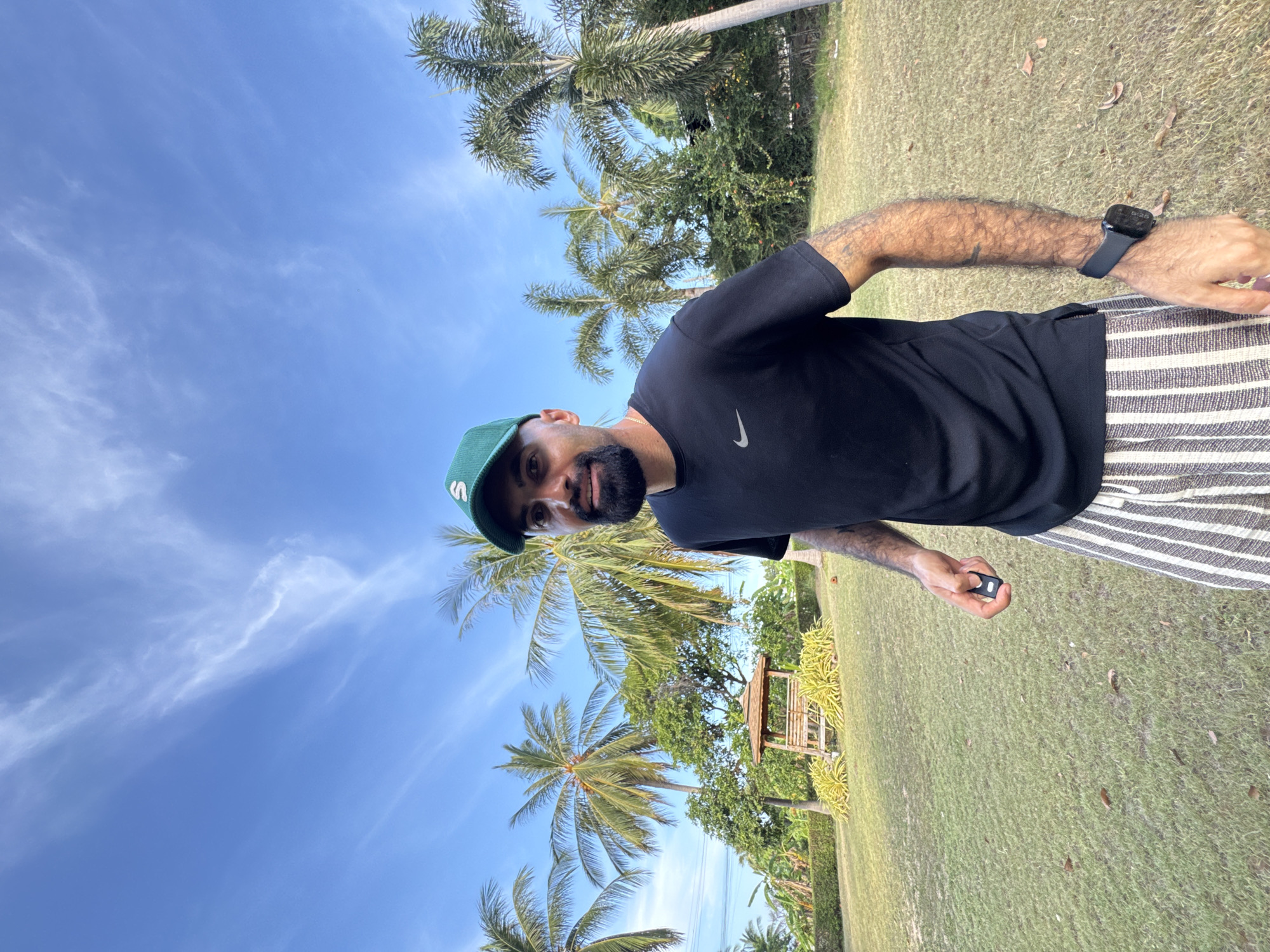 A man wearing a green baseball cap with a white "S" logo, a black t-shirt with a Nike logo, and striped pants stands in a grassy yard with palm trees and a gazebo in the background. The sky is blue with wispy white clouds. The man is holding a small black object in his right hand and wearing a black smartwatch on his left wrist. The scene appears to be outdoors in a tropical location, likely Ko Samui District, Thailand.