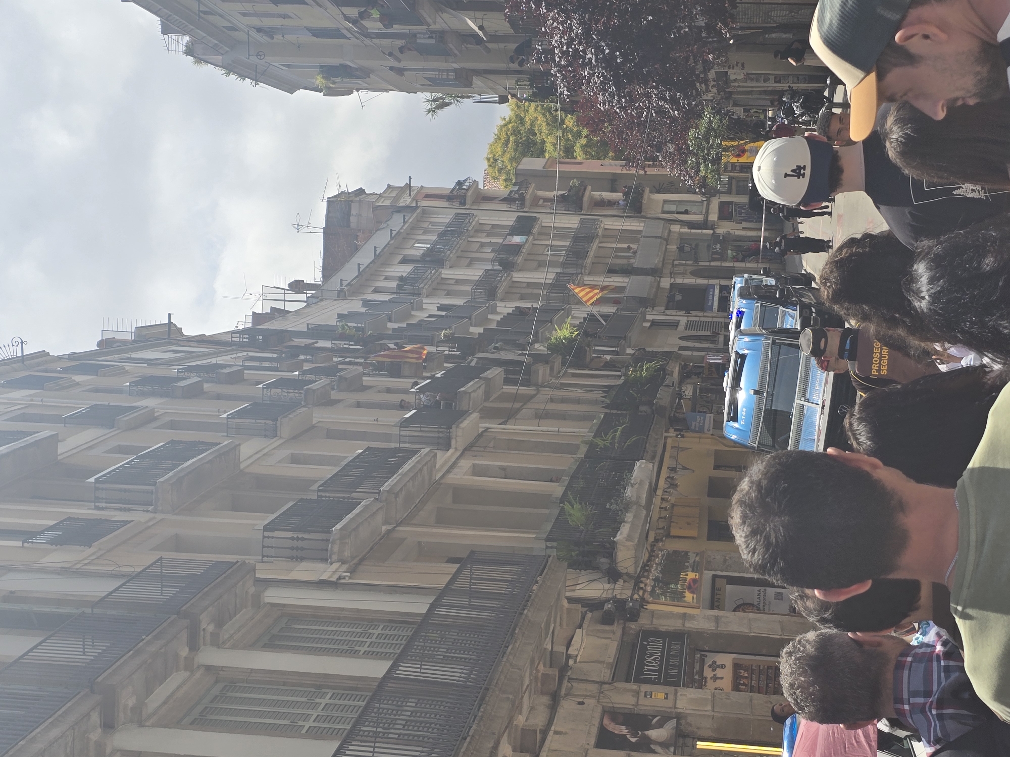 The image depicts a street scene in Barcelona, Spain, characterized by tall, narrow buildings with balconies and shutters lining both sides of a pedestrian street. Several people are visible in the foreground, facing away from the camera, suggesting they are observing something down the street. In the middle ground, a police presence is evident with at least two blue police vans and several uniformed officers. There are also red and white barrier tape across the street, indicating a secured or cordoned-off area. Flags, possibly regional or political, are displayed on some balconies. The overall atmosphere appears to be one of public gathering or a potentially tense situation, given the police activity. The architecture suggests a historic European city, and the presence of Catalan flags (if they are indeed Catalan flags) would further point to Barcelona.