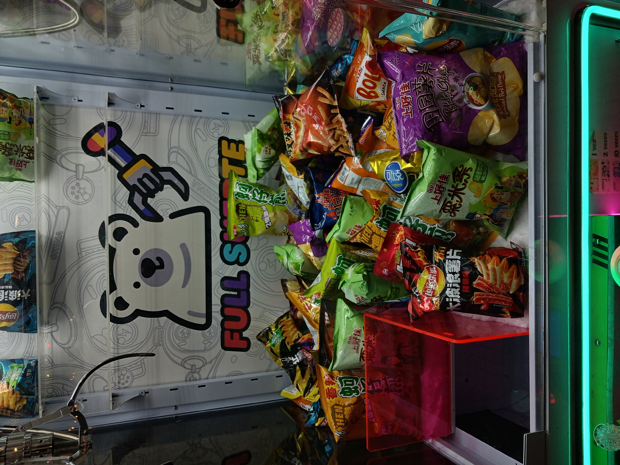 This media file captures the brightly lit interior of a claw game machine, abundantly filled with numerous bags of chips and other snacks. The scene is set indoors, likely within an arcade or entertainment venue, characterized by the machine's design and neon lighting. The context provided specifies the location as Buenos Aires, Argentina.
The machine's contents are predominantly snack foods, with a visible assortment of potato chips and other packaged treats. Many items feature East Asian branding and flavors. Notable brands include Lay's (乐事), Oishi (上好佳), Copico (可比克), and BoL snacks. Specific product details and flavors indicated on the packaging include "马铃薯片" (Potato chips), "大波浪" (Big Wave) chips, "铁板" (Teppanyaki/Hot Plate) flavor, "粟米条" (Corn sticks), "哈密瓜味" (Hami Melon flavor), "香辣" (Spicy), "蚵仔煎" (Oyster Omelet) flavor, "辣条" (Spicy strips), and "Braised Pork Flavor" (卤肉口味). Some packaging also features the word "CHINA" and "2倍濃" (2x concentrated).
A metallic, three-pronged claw mechanism is visible in the lower left, positioned above the pile of snacks. The machine's back panel displays a white graphic background with outlines of gears and chains, featuring a cartoon bear character and the colorful text "FULL SQUARE." A robotic arm graphic is also part of this internal design. The right side of the machine is illuminated by vibrant green and pink neon lights. Adjacent to these lights, text in Spanish reads "1 vez," "2 veces," and "10 veces" (1 time, 2 times, 10 times), likely indicating options for gameplay attempts or credits. The overall presentation is vivid and engaging, typical of arcade prize machines.