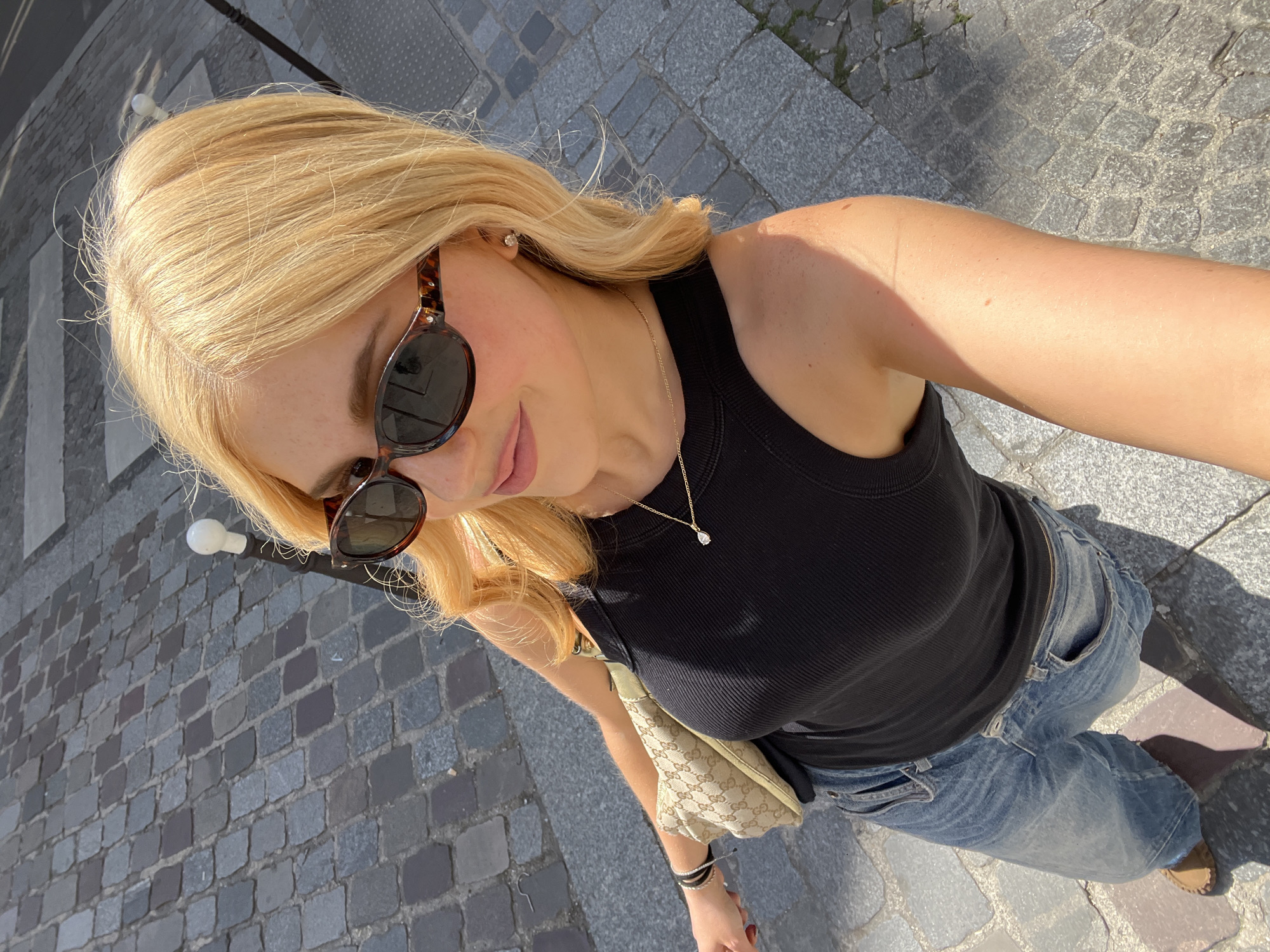 A young, fair-skinned woman with blonde hair is taking a selfie in an urban setting, likely in Paris, France, based on the cobblestone pavement. She is wearing sunglasses, a black tank top, and blue jeans. A beige Gucci bag is slung across her body. She also wears a delicate necklace and bracelets on her wrist. The pavement consists of small, irregularly shaped cobblestones. A small lamppost is seen in the background. The lighting suggests it is daytime, and the sun is shining.