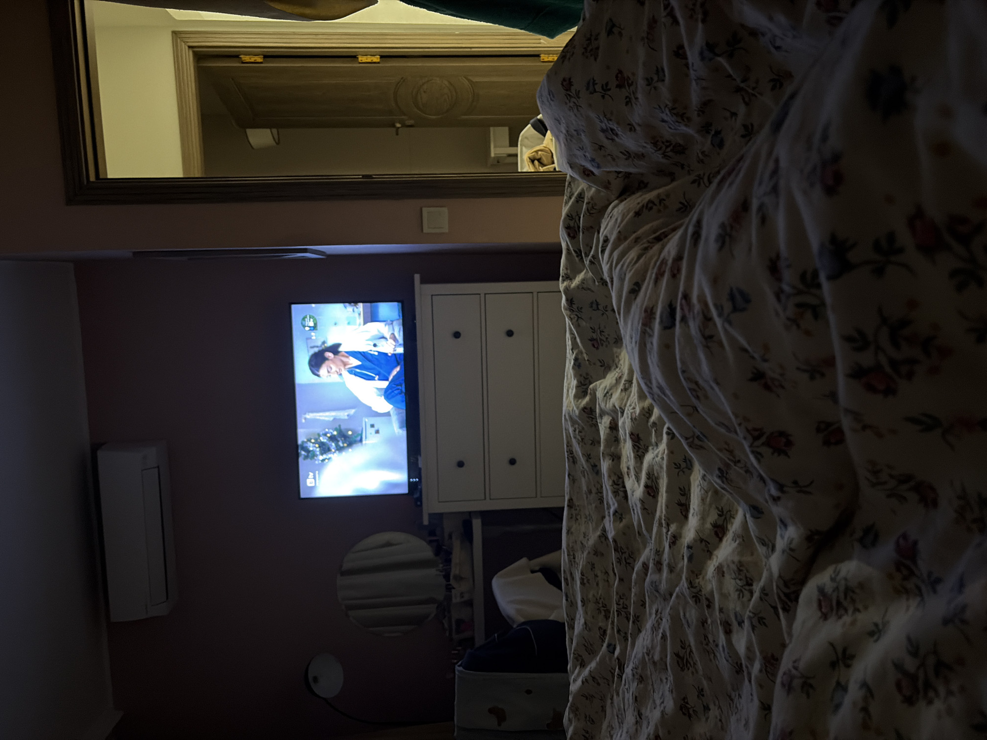 The image shows a bedroom with a bed covered in a floral patterned comforter in the foreground. In the background, there is a pink wall with a television displaying a scene of a woman in scrubs with a Christmas tree in the background. To the right of the TV is a white cabinet. Below the TV is a small round mirror and an air conditioner unit. Above the TV, there is a mirror with a wooden frame reflecting a room. There is no indication that this scene is located in Burgas, Bulgaria.