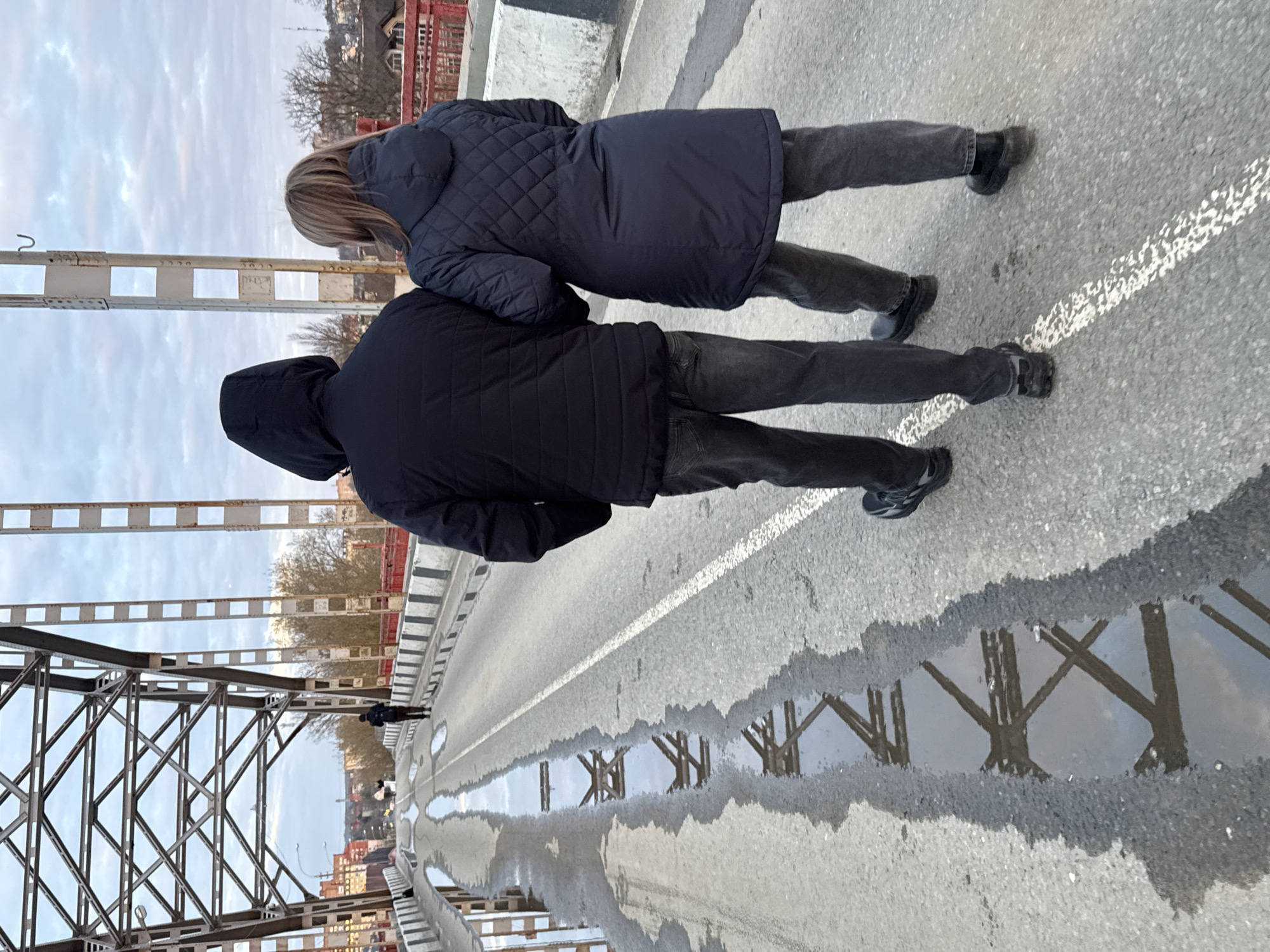 The image, captured in Dmitrov, Russia, depicts two individuals walking on a bridge on an overcast day. The scene is viewed from behind the subjects, who are positioned towards the center-right of the frame, moving away from the viewer.
In the foreground, one person wears a dark hooded jacket and dark pants. Slightly ahead and to their right, another person with blonde hair is seen from the back, wearing a dark, quilted winter coat and dark pants. Both appear to be casually traversing the bridge.
To the left, the prominent metal truss structure of the bridge is visible, with its intersecting beams creating geometric patterns. The road surface is asphalt, featuring a faded white dashed line. Scattered across the road are numerous puddles, which reflect the intricate angular forms of the bridge's metal trusses, creating striking visual echoes. In the background, beyond the bridge structure, a distant cityscape with various buildings and trees can be observed under the muted light of the cloudy sky. Small white rectangular signs are attached to the bridge's metal beams; some display partially visible numbers, such as "1220" and "2330", though they are mostly indistinct. The presence of puddles suggests recent rain or melting conditions, contributing to the cool and muted atmosphere of the scene.
