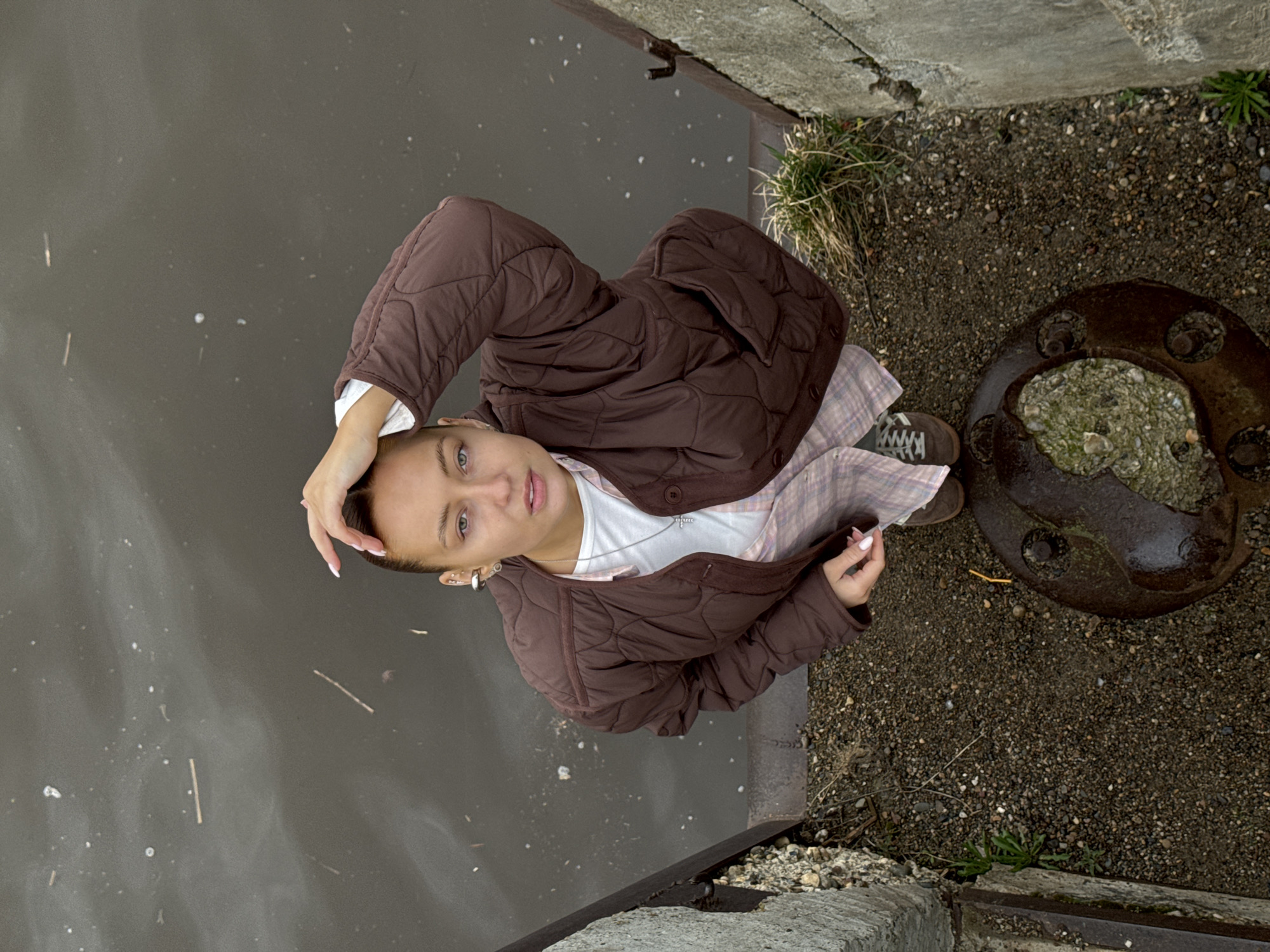 A young woman stands near a body of water in Chastyye, Russia. She has short hair and wears a brown quilted jacket over a white shirt and patterned dress. Her left hand is raised to her head, while her right hand holds an object. She stands on gravel near a concrete structure and a rusty metal object. The water appears murky and contains debris.