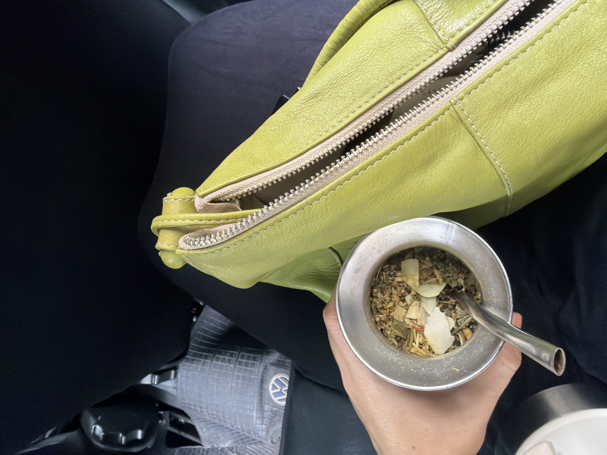 A person is sitting in the passenger seat of a vehicle. They are holding a mate gourd filled with yerba mate and a bombilla in their hand. A green leather purse is next to the mate. The vehicle has a Volkswagen emblem on the seat. The location is likely Buenos Aires, Argentina due to the mate, which is a common drink there.