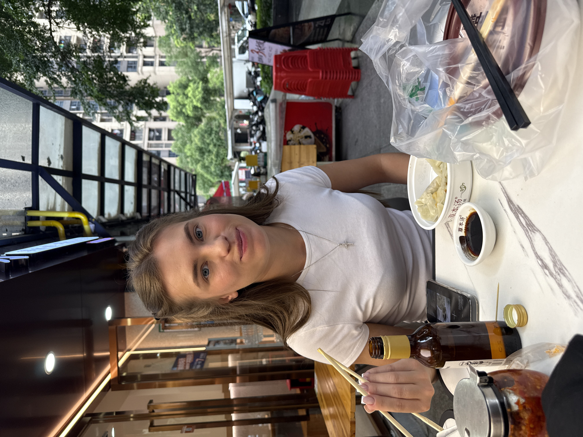 A young woman with blonde hair pulled back and blue eyes smiles at the camera, holding a pair of chopsticks. She is seated at a table outdoors, likely at a restaurant in Haizhu District, China, as indicated by the architectural style and surrounding environment, including a building with multiple stories and trees. The table is covered with a marble-patterned tablecloth and set with a bowl of dumplings, a small dish of soy sauce, a bottle of amber liquid with a yellow cap, a phone, a jar of red chili paste, and a plastic bag with chopsticks and possibly another dish inside. In the background, there is a covered walkway with yellow pipes and modern buildings, as well as parked motorcycles and scooters. Red plastic chairs are stacked nearby, and an A-frame sign is visible. The overall scene suggests a casual meal outdoors in an urban setting.
