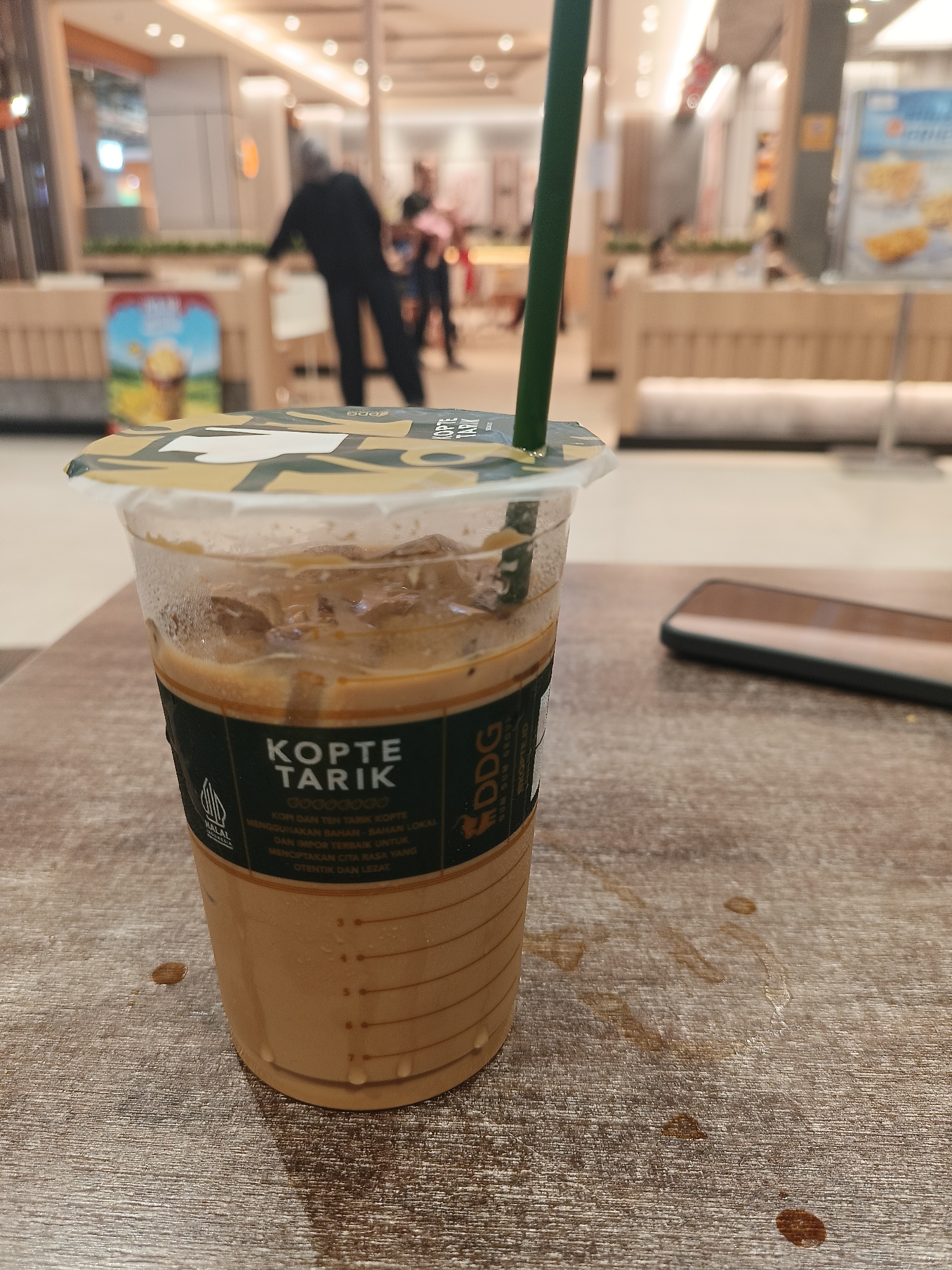 A close-up, eye-level shot focuses on a plastic cup of iced "Kopte Tarik" coffee placed on a wooden table. The cup, which has condensation on its exterior, is about two-thirds full with a light brown liquid and ice cubes. A green straw extends from the cup's partially opened lid. A dark green label wraps around the lower half of the cup, displaying "KOPTE TARIK" in white text, along with smaller text in white and gold, and a "HALAL" certification logo. The table surface is textured and shows several small, dark brown droplets, possibly from spilled coffee. In the blurred background, people can be seen in what appears to be a retail or food court environment. The overall lighting suggests an indoor setting. There is no specific visual information in the image to indicate the location in Palangka Raya, Indonesia.