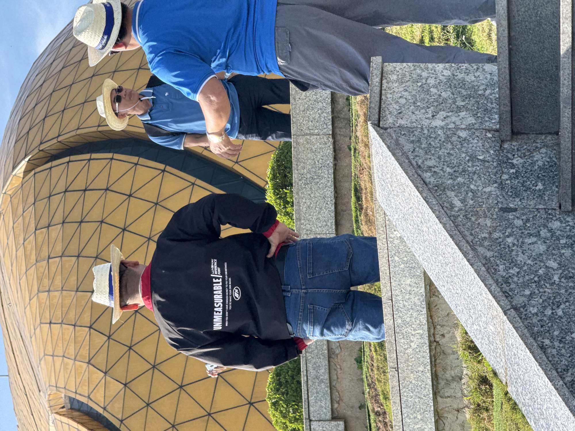 Three men are standing in front of a large golden dome structure in Da Lat, Vietnam. The dome is covered in a geometric pattern of triangles and appears to be a prominent architectural landmark. The man in the foreground, facing away from the camera, wears a black jacket with "UNMEASURABLE" printed in white text on the back, blue jeans, and a straw hat. He is standing on a set of granite stairs. To his right, another man in a blue polo shirt and dark pants also wears a straw hat and sunglasses. He is speaking to the third man, who is wearing a blue polo shirt with white trim, grey pants, and a straw hat. The third man is partially obscured from the front but is facing the man in the blue polo shirt, with his right hand extended as if gesturing. The environment appears to be outdoors, with lush green grass and trees visible behind the men and the dome. The sky is clear and blue.