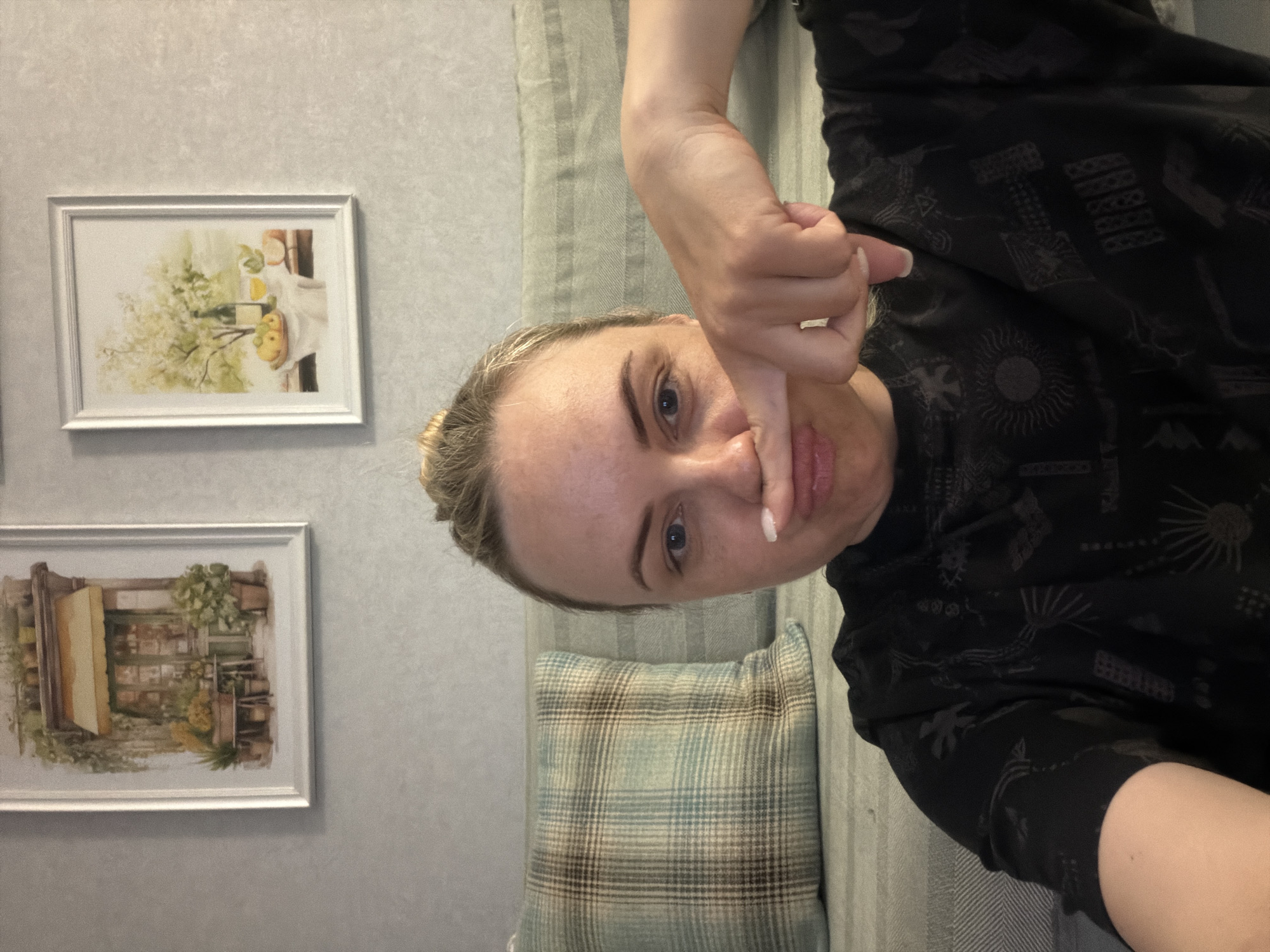 A woman is lying on a couch. She is wearing a black shirt with a pattern. She is holding her right hand up with her thumb pointing down. Behind the woman is a couch with a striped pattern and a plaid pillow. On the wall are two framed prints. The location cannot be verified as Votkinsk, Russia based on the image provided.