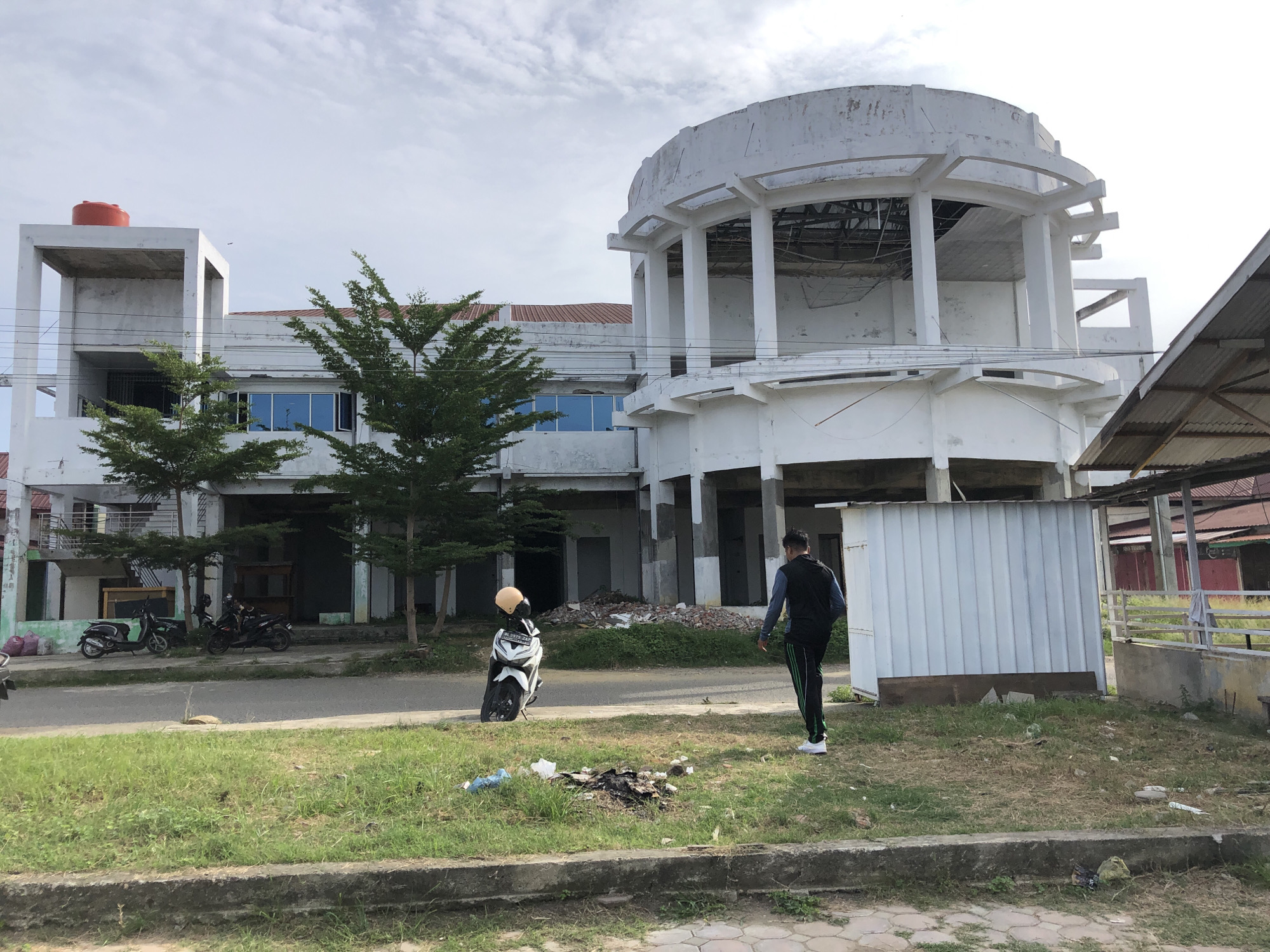 In the city of Lhokseumawe, Indonesia, a partially constructed building stands under a cloudy sky. The structure features a prominent circular section with visible scaffolding and an open roof, suggesting ongoing construction or renovation. To the left, a lower, rectangular building with a red water tank on its rooftop is attached. The surrounding area is paved with grass and a sidewalk, with debris scattered on the ground. Two motorcycles are parked near the building's entrance, and a lone person, wearing a dark vest over a light blue long-sleeved shirt and black pants with green stripes, walks away from the camera towards the right side of the frame. Another motorcycle, white and black, is parked to the left of the walking individual, with a yellow helmet resting on its seat.