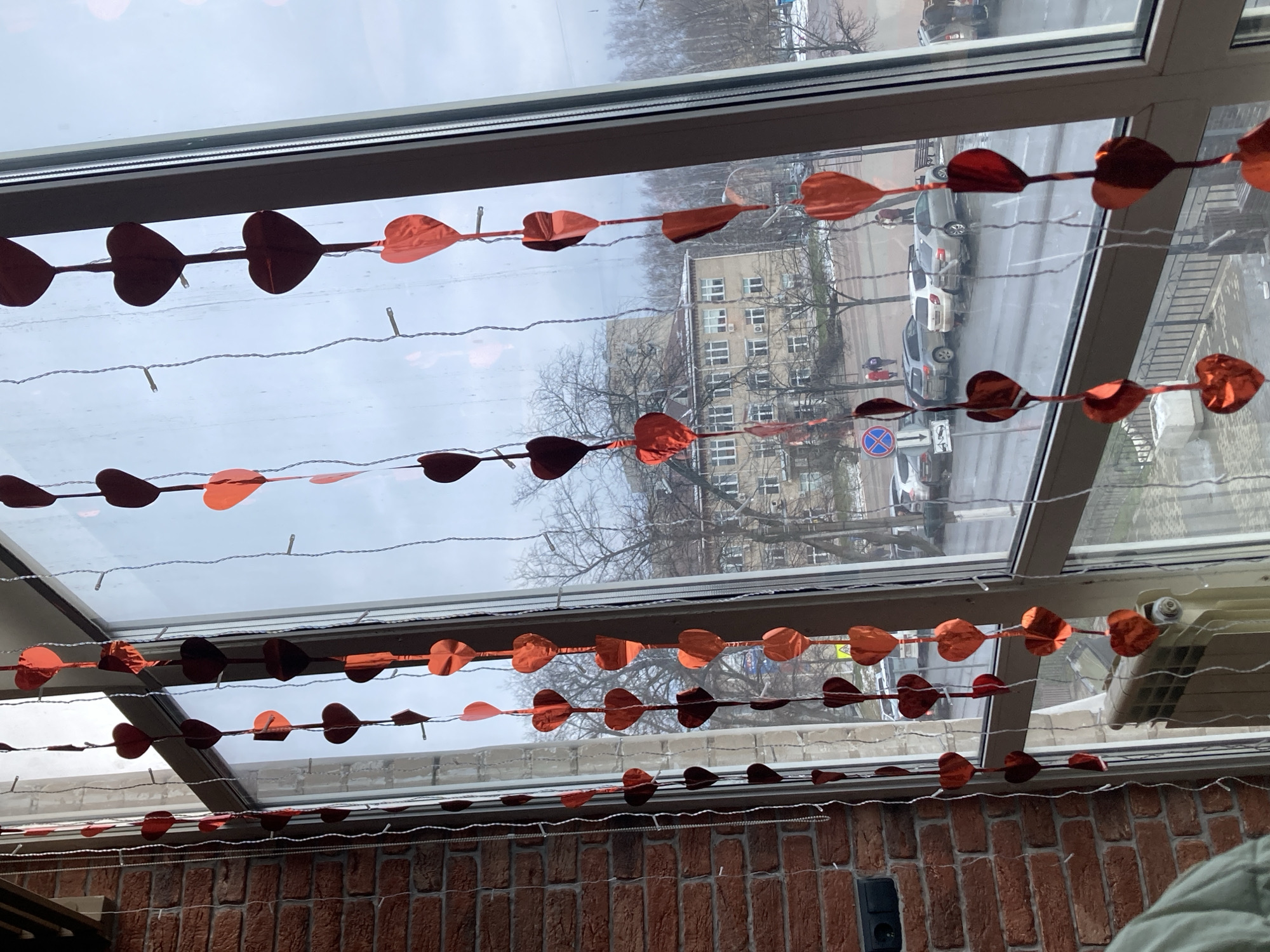 A photograph taken indoors looking through a window shows a brick wall at the bottom, and a series of strings of red heart decorations hanging across the window. Outside, there are buildings, trees, a street, and some parked cars. The image suggests an urban setting on an overcast day. This is a domestic scene, as the decorations and the presence of a brick wall suggest an indoor space, decorated perhaps for Valentine's Day. The location is Chaikovsky, Russia.