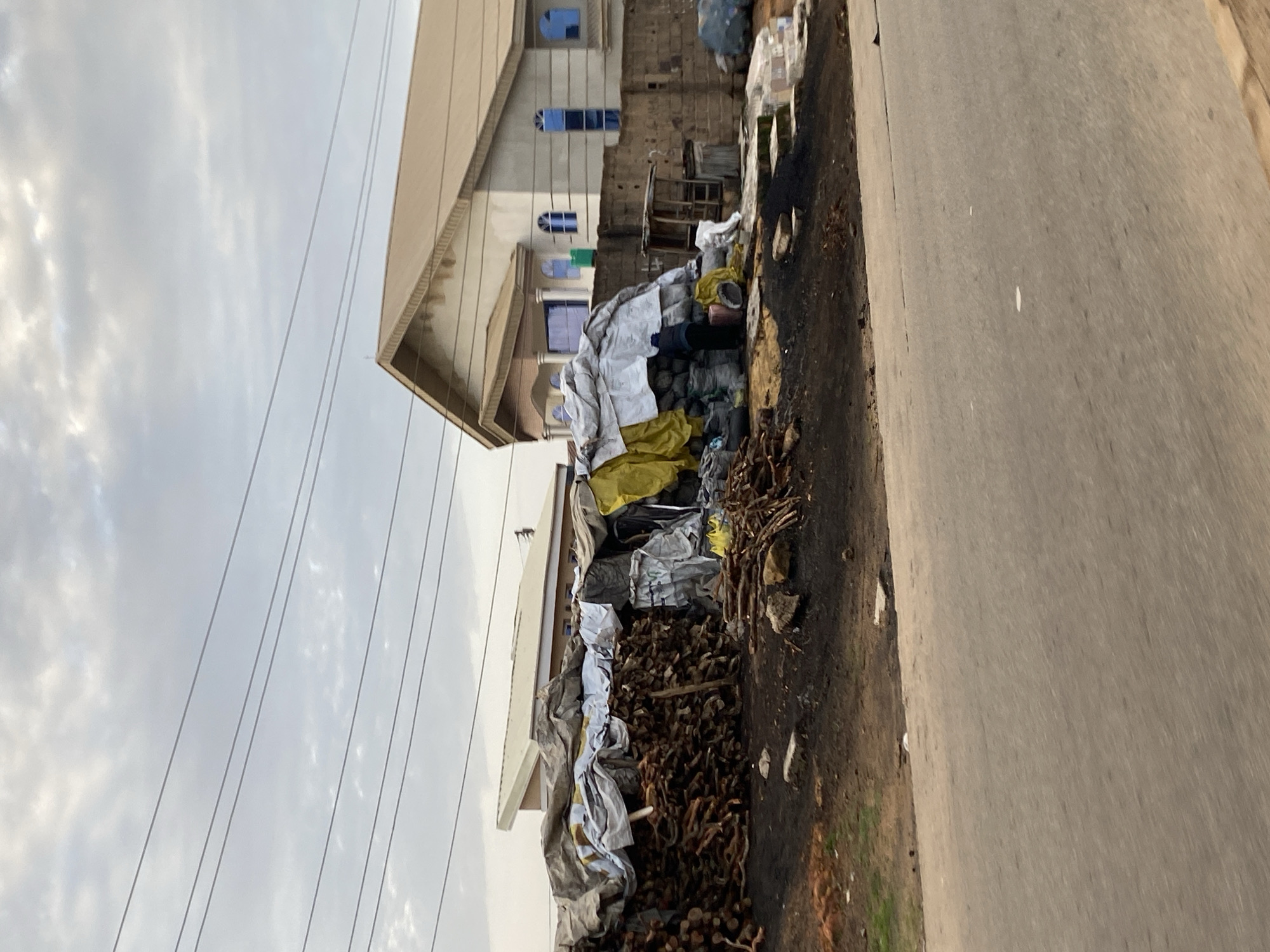 In Barakin Lerawi, Nigeria, a scene unfolds along a paved road. A substantial pile of chopped wood, covered partly by tarpaulins and yellow sheeting, occupies the foreground. The wood is arranged in a dense stack, with some pieces scattered around its base. Behind the wood pile, a brick wall encloses a yard. A small, open-sided wooden shelter with a corrugated roof stands against the wall. Within the yard, more assorted items are piled, some covered with sheeting, and sacks are visible. A person, dressed in dark clothing, stands near the sacks, facing away from the viewer. To the right of the wall, a building with a light-colored, peaked roof and blue-tinted windows is visible. Power lines crisscross the sky, which is overcast with clouds. The ground in front of the woodpile appears to be dark soil or ash.