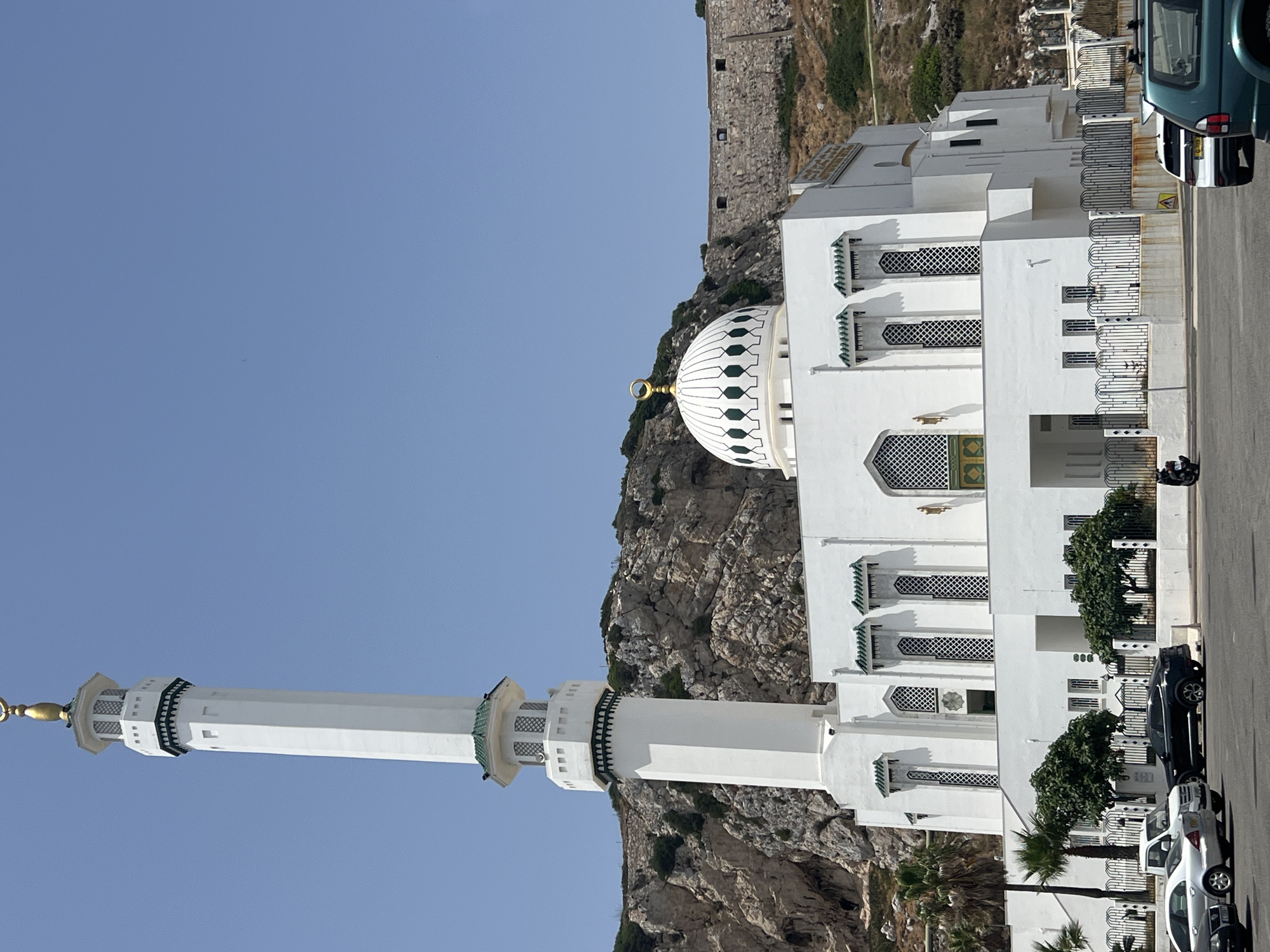 The image shows the Ibrahim-al-Ibrahim Mosque, located in Europa Point, Gibraltar. The mosque is a white building with a tall, slender minaret and a prominent dome topped with a golden crescent. It is set against a backdrop of a steep, rocky hillside, part of the Rock of Gibraltar, with a historic stone wall visible further up the slope. In the foreground, there is a parking area with several cars parked, including silver sedans and a dark SUV. Some trees and palm trees are also visible around the mosque. The sky is clear and blue.