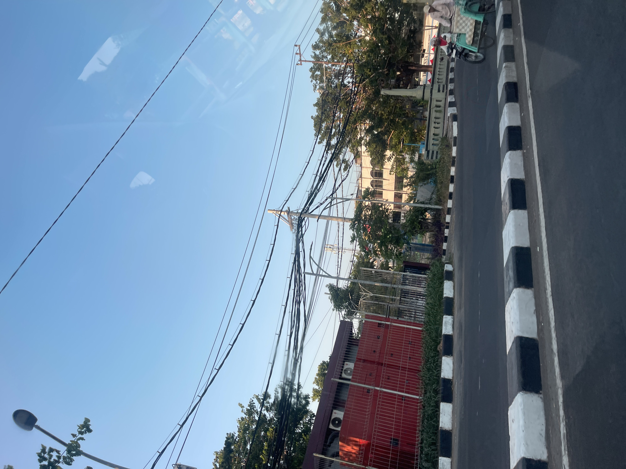 This is an eye-level, medium shot captured from a vehicle, likely a car, looking towards a city street in Surabaya, Indonesia. The sky is a clear, bright blue with no clouds. Prominently featured are numerous overhead electrical wires, creating a dense, tangled network. A street lamp is visible on the left, and a large red building with visible air conditioning units is behind a metal fence. Trees with green foliage are scattered throughout the scene, with some closer to the foreground and others further back. The road has distinct black and white striped bollards lining the side. In the middle ground, a man wearing a white cap and light-colored shirt is riding a motorcycle with a sidecar, which appears to be carrying goods or passengers covered with a checkered blanket. The building behind the motorcycle is partially visible and seems to be a commercial or public building with a sign partially obscured but readable as "TRANSA". The overall impression is a typical urban street scene in Surabaya under a clear sky.
