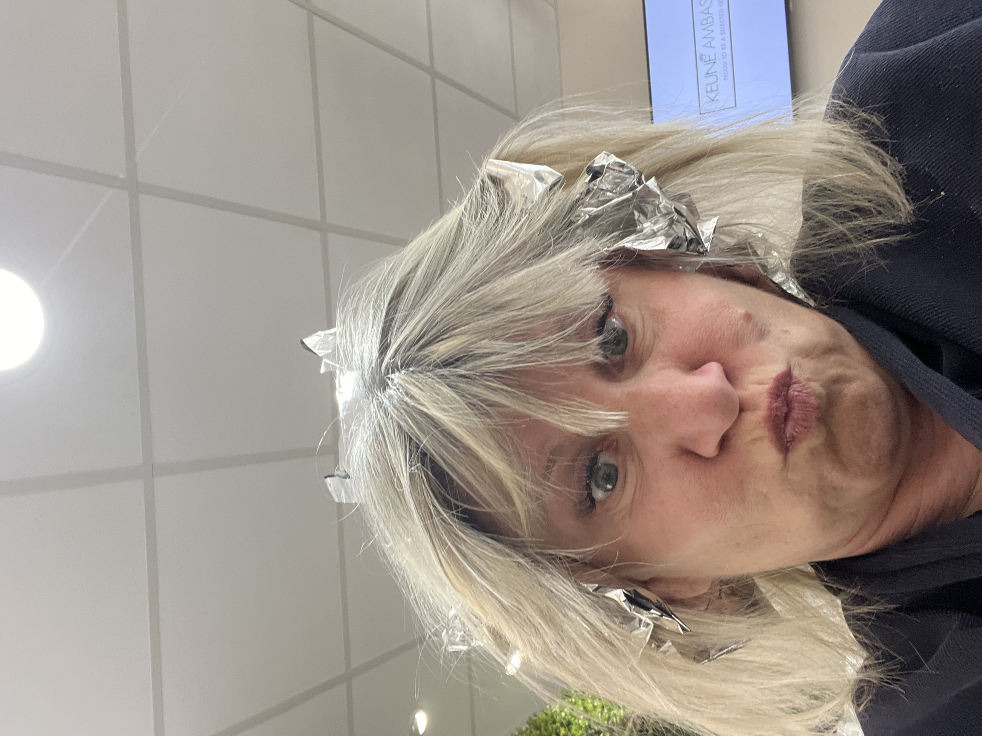 A woman with blonde hair and foil highlights is puckering her lips, seemingly taking a selfie indoors. The background features a grid-patterned white wall, suggesting a salon setting. A sign with the word "KEUNE AMBAS" is partially visible. A light fixture is present in the background. The location, Montrond-les-Bains, France, could be associated with the salon in the picture.