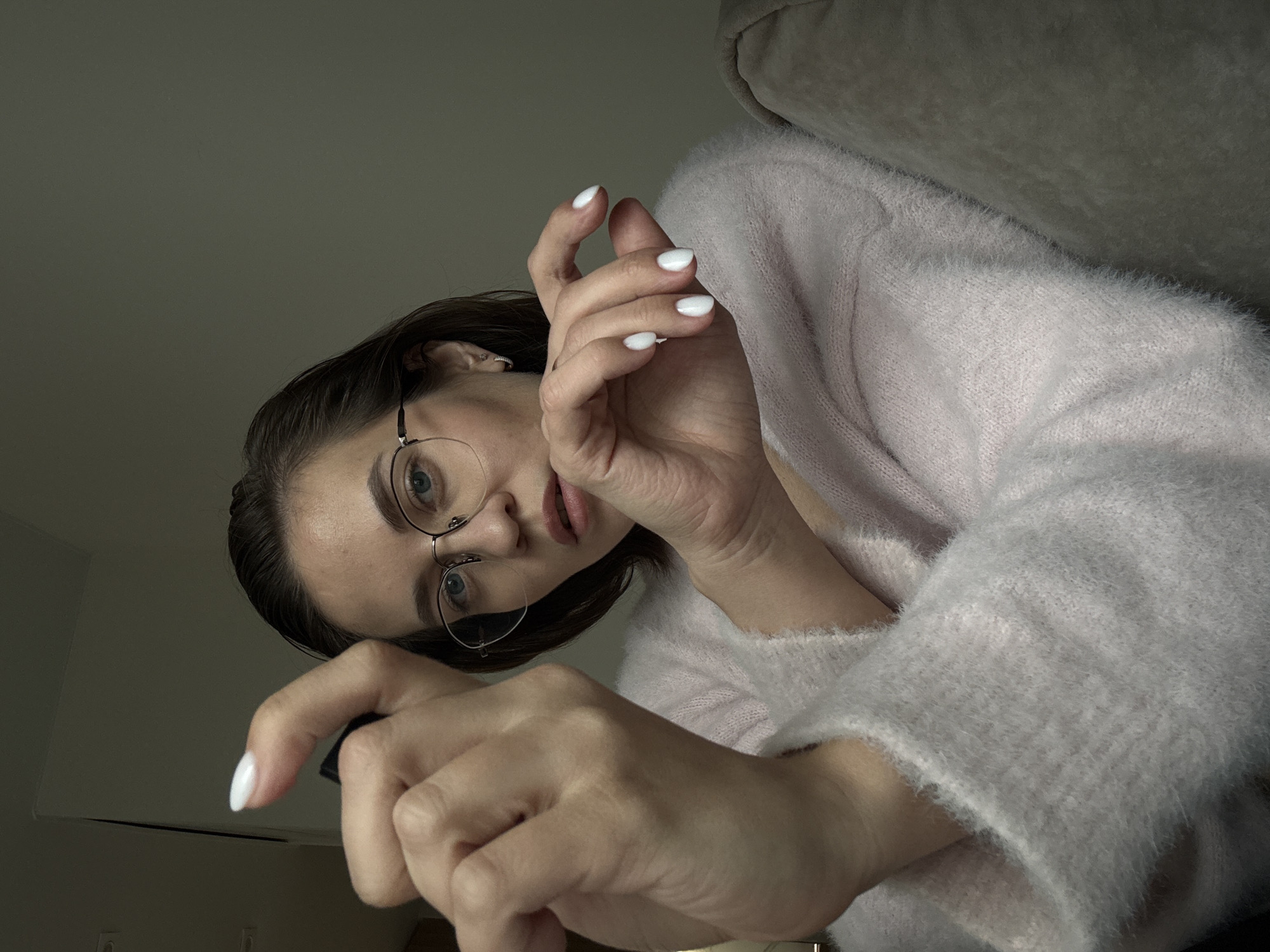 A woman with short brown hair, wearing rimless glasses and a light pink, fuzzy sweater, is positioned at an unusual angle, looking down at the viewer. Her nails are painted white. She is holding a dark object in her left hand. The background consists of a light-colored wall and a gray surface, potentially part of a sofa or other furniture. The lighting is dim. The location is unknown.