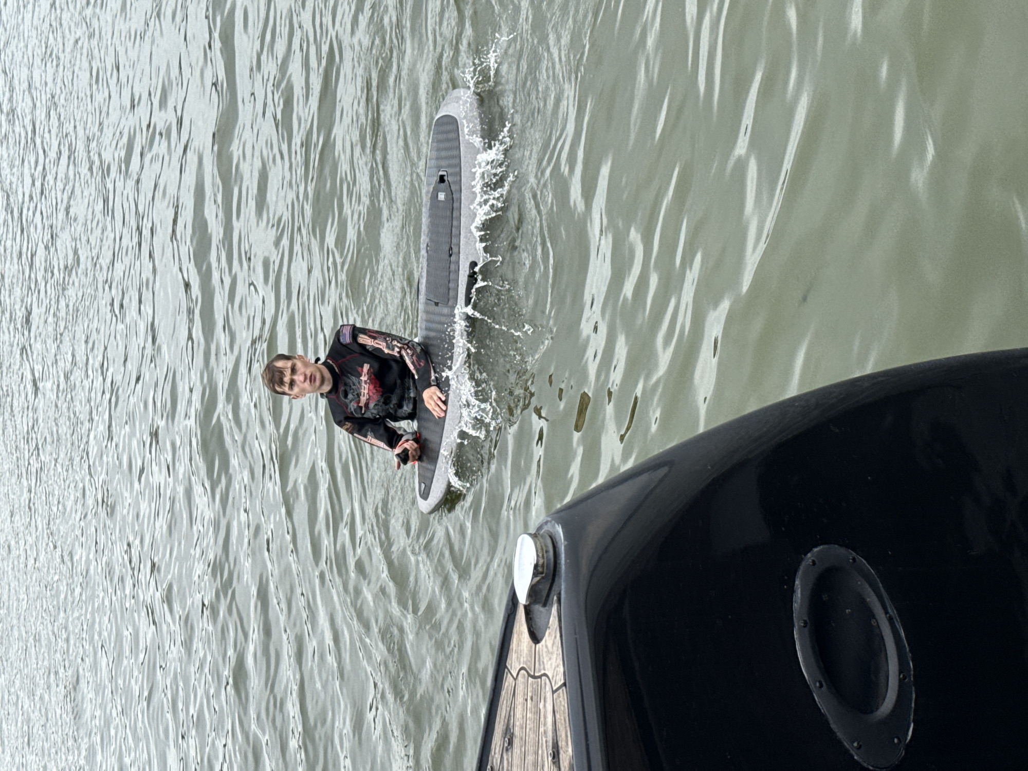 In Votkinsk, Russia, a young man wearing a black and red wetsuit is sitting on a motorized surfboard on the surface of a body of water. He is holding a black remote control in his right hand and his left hand rests on the surfboard. The surfboard is partially submerged, creating a small splash. In the foreground, the stern of a black boat is visible, with a wooden deck and a metal cleat. The water is choppy, with ripples and waves. The sky is overcast.