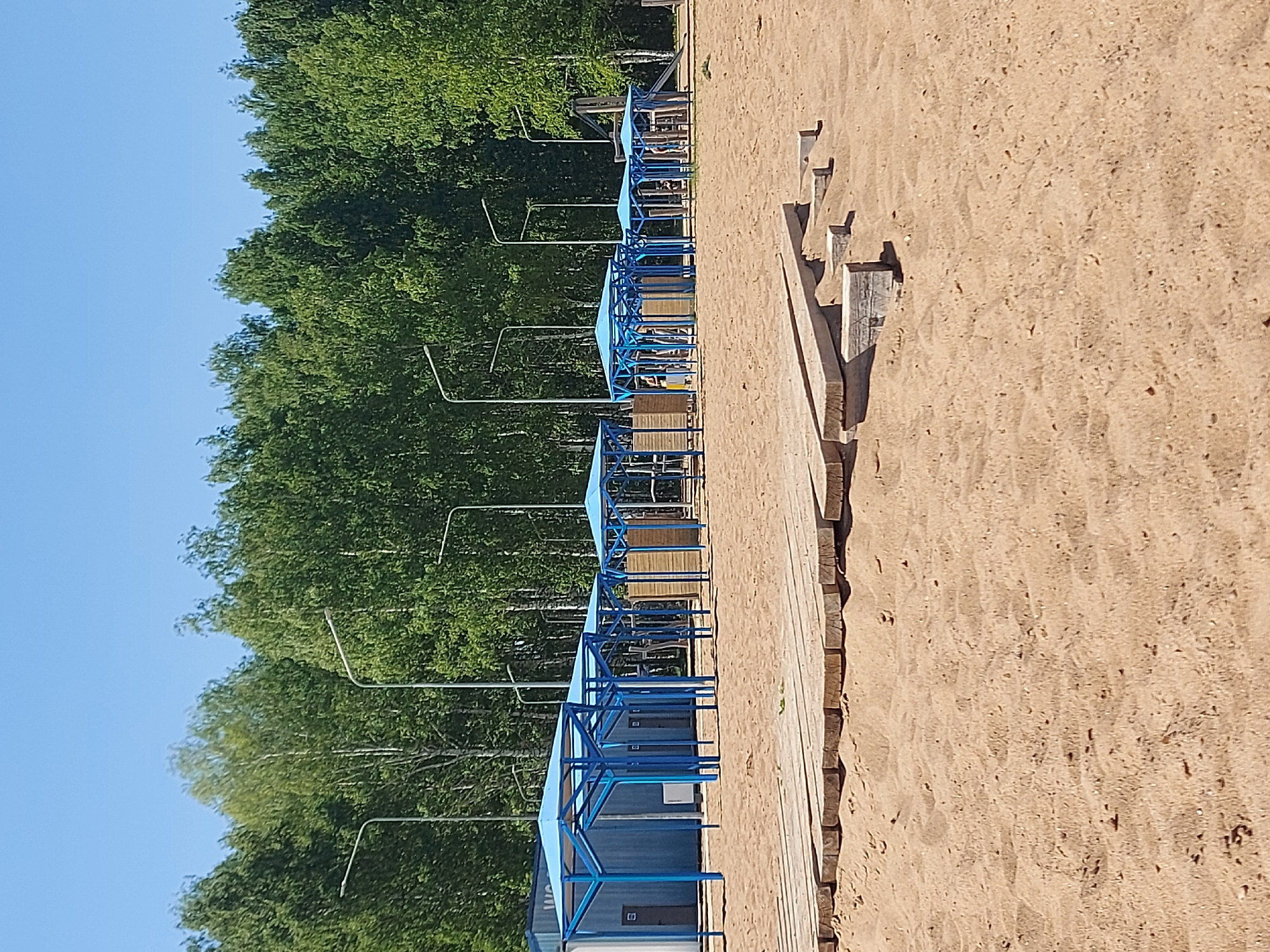 The image depicts a beach scene in what is likely Chaikovsky, Russia, characterized by light-colored sand. A line of blue cabanas or changing stalls extends into the distance, positioned adjacent to a wooden walkway. Tall green trees form a backdrop to the beach structures, and streetlights are visible interspersed among the trees. The sky is clear and blue, suggesting a sunny day. No individuals are clearly visible in the immediate foreground, but there appear to be people occupying the cabanas. The photograph's orientation is vertical.