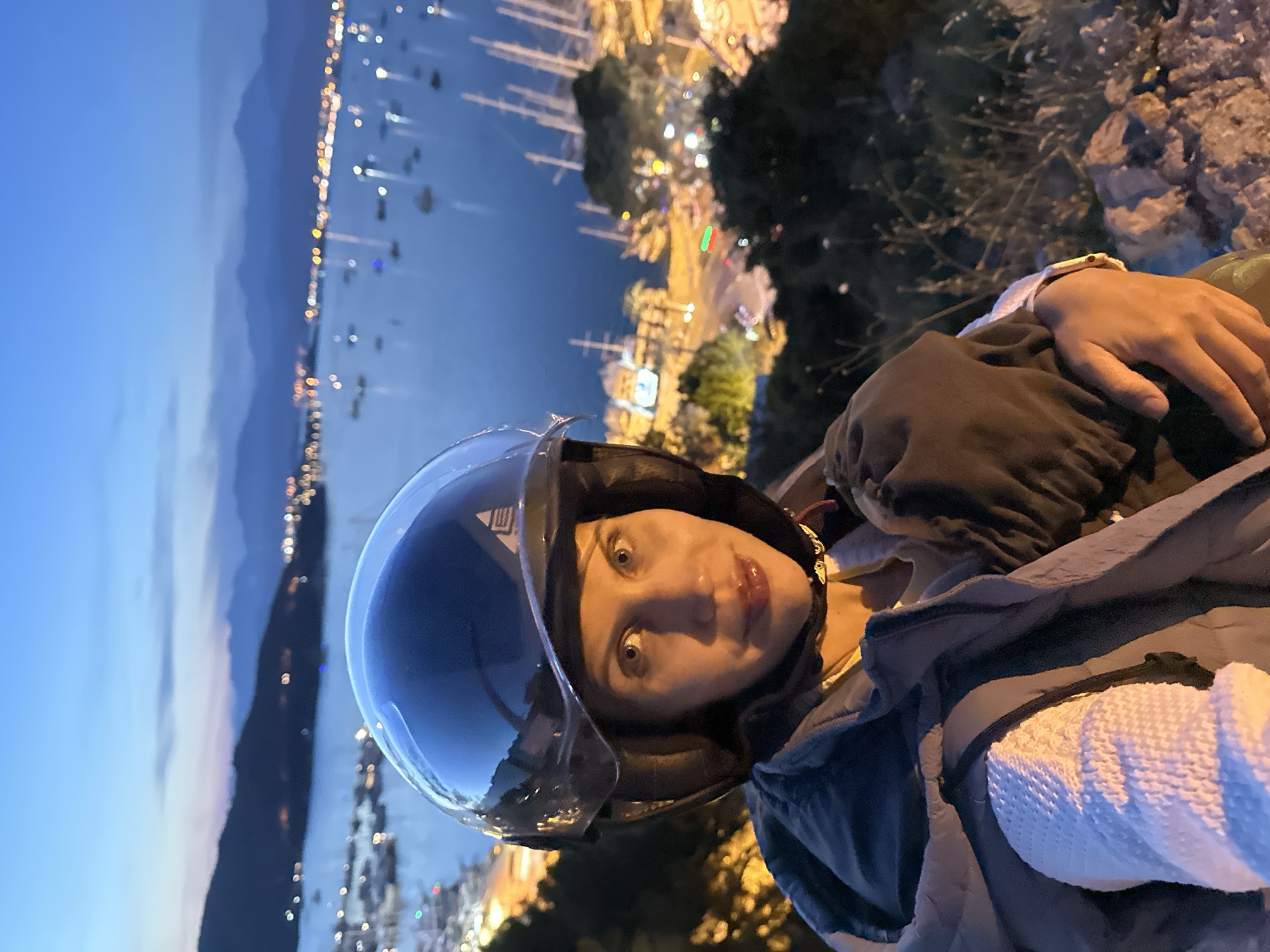 A person wearing a motorcycle helmet is positioned at an elevated location overlooking the city of Fethiye, Turkey at night. The cityscape and harbor are visible in the background, dotted with lights. The person is wearing a jacket and possibly bundled in a blanket. The environment appears to be a rocky hillside or overlook point.