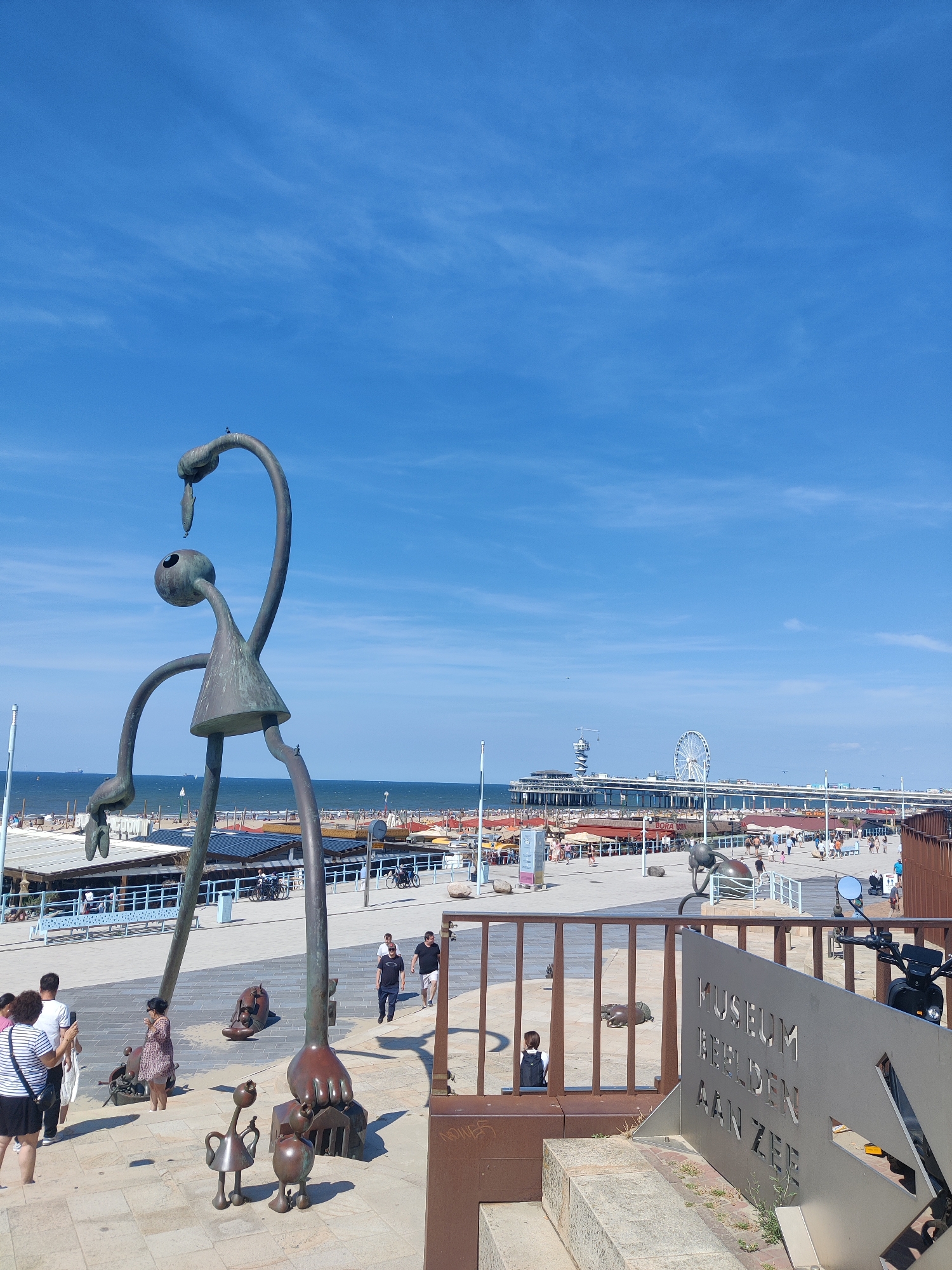 This is a full-frame, eye-level, slightly elevated shot of a seaside promenade in Scheveningen, The Hague, Netherlands. In the foreground, to the left, is a large, abstract bronze sculpture depicting a stylized human-like figure with elongated limbs, a triangular skirt, and a rounded head with a single eye. The sculpture's neck curves upwards, and its head is tilted as if interacting with something small perched on its neck. Smaller bronze sculptures of stylized figures and abstract shapes are scattered around its base.
Behind the sculptures, a paved promenade stretches towards the sea. People are walking and standing on the promenade. Beach umbrellas and structures are visible on the sandy beach beyond the promenade. In the distance, a long pier extends into the ocean, featuring a Ferris wheel and a tall tower structure. The sea is visible behind the pier, with several boats on the water. The sky is a clear, bright blue with a few wispy white clouds.
To the right of the main sculpture, a metal sign with embossed lettering reads "MUSEUM BEELDEN AAN ZEE." A black scooter is parked near the sign. The overall scene depicts a sunny day at a popular tourist destination by the sea.